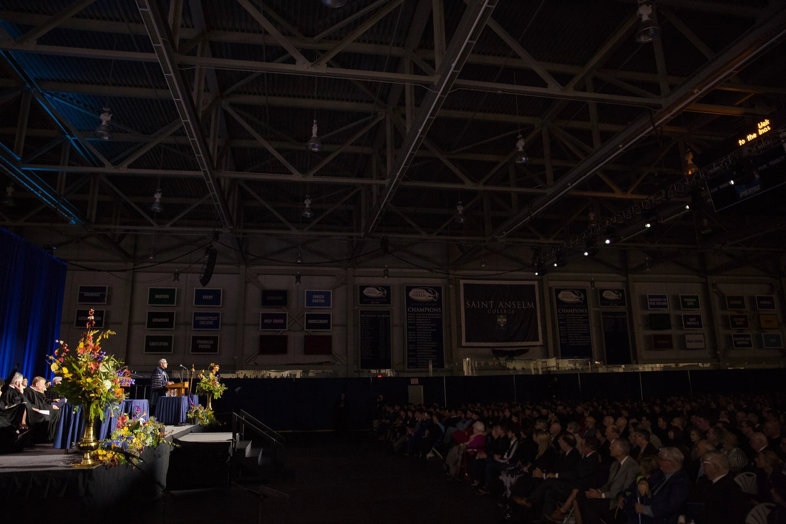 An indoor graduation ceremony at Saint Anselm College with students receiving diplomas on stage and an audience watching in the darkened auditorium.