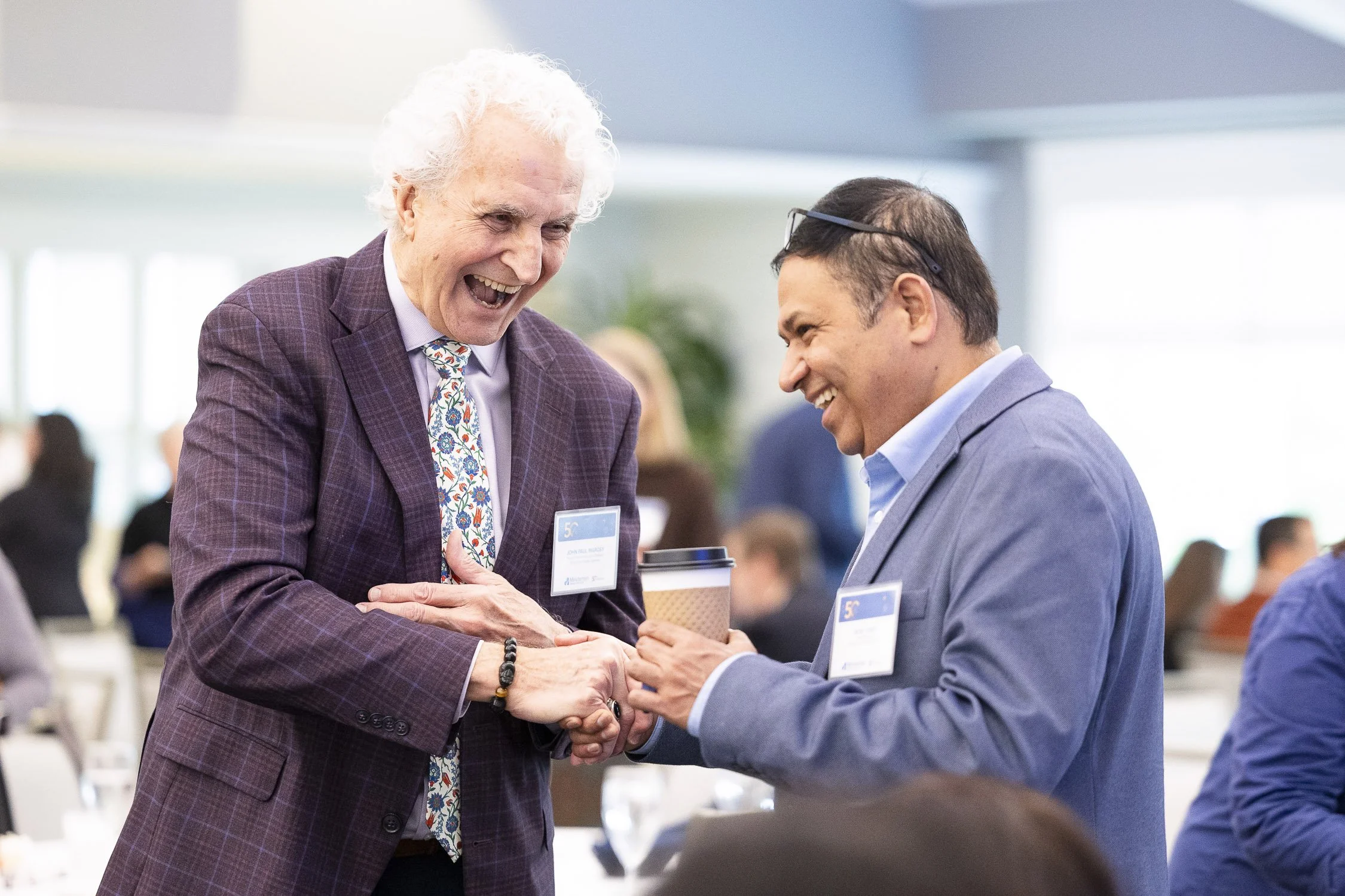 Two men, are smiling and shaking hands at a nonprofit luncheon event in Lincoln, Massachusetts.