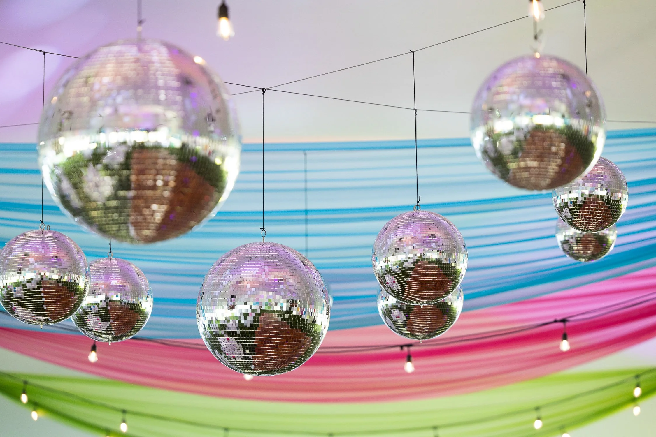 Multiple disco balls hanging from the ceiling with colorful fabric drapes in the background and string lights.