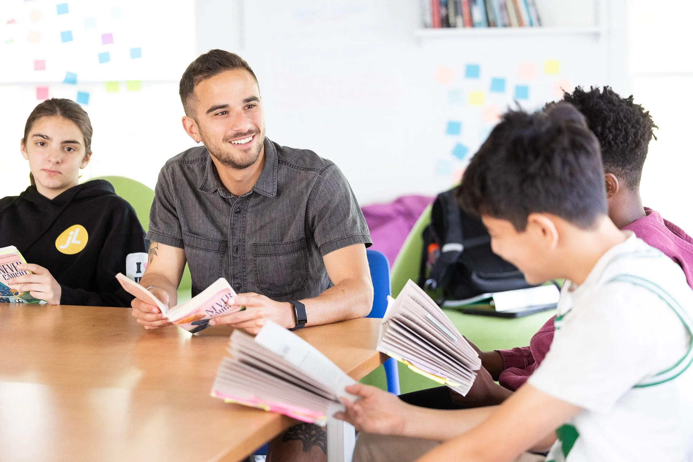 A teacher at independent school sitting at a classroom table with three students, smiling and holding books, in a bright classroom with colorful sticky notes on the wall.