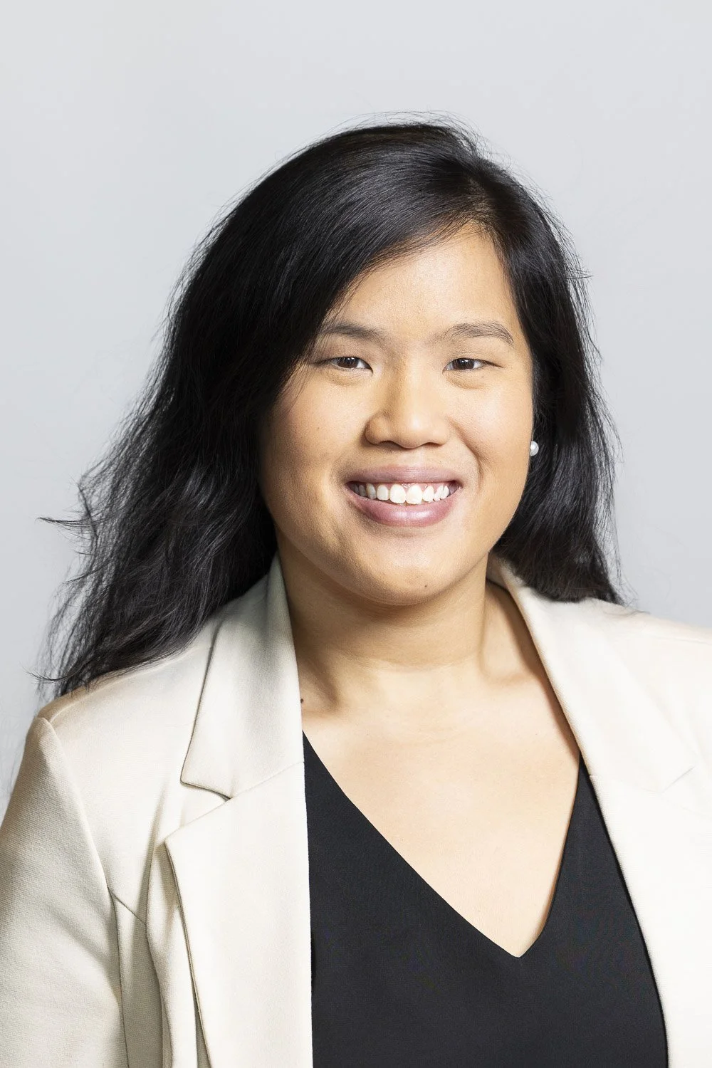 Professional headshot of a woman with long black hair, wearing a light-colored blazer and black top, against a plain light gray background.