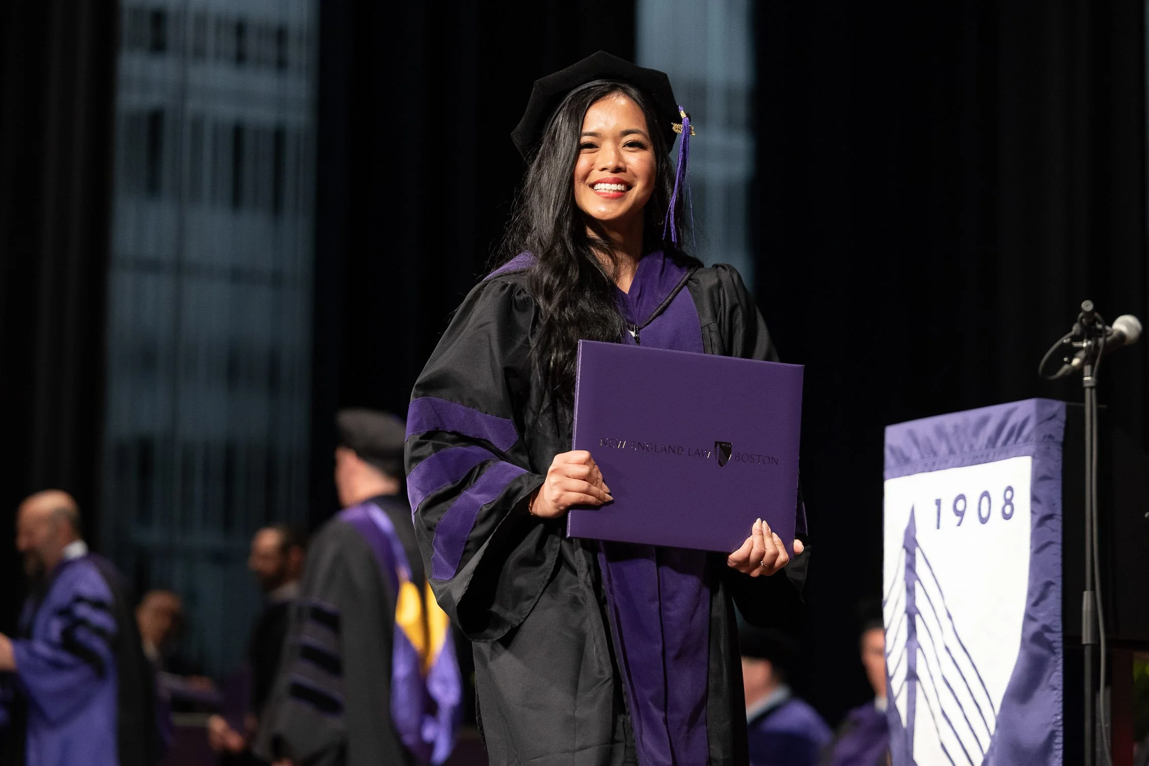 A woman dressed in graduation regalia smiling and holding a diploma on stage during a graduation ceremony at New England Law Boston.