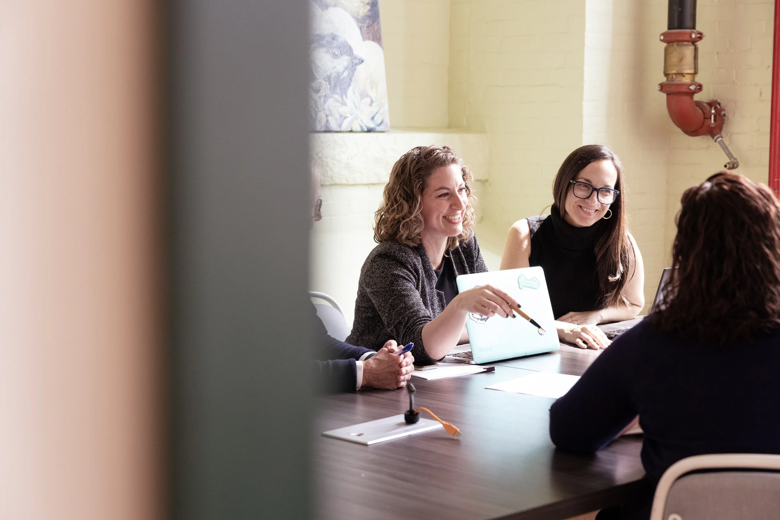 Four women sitting around a conference table, smiling and chatting during a meeting in a bright room. One woman has a laptop decorated with stickers, and another has glasses and dark hair.