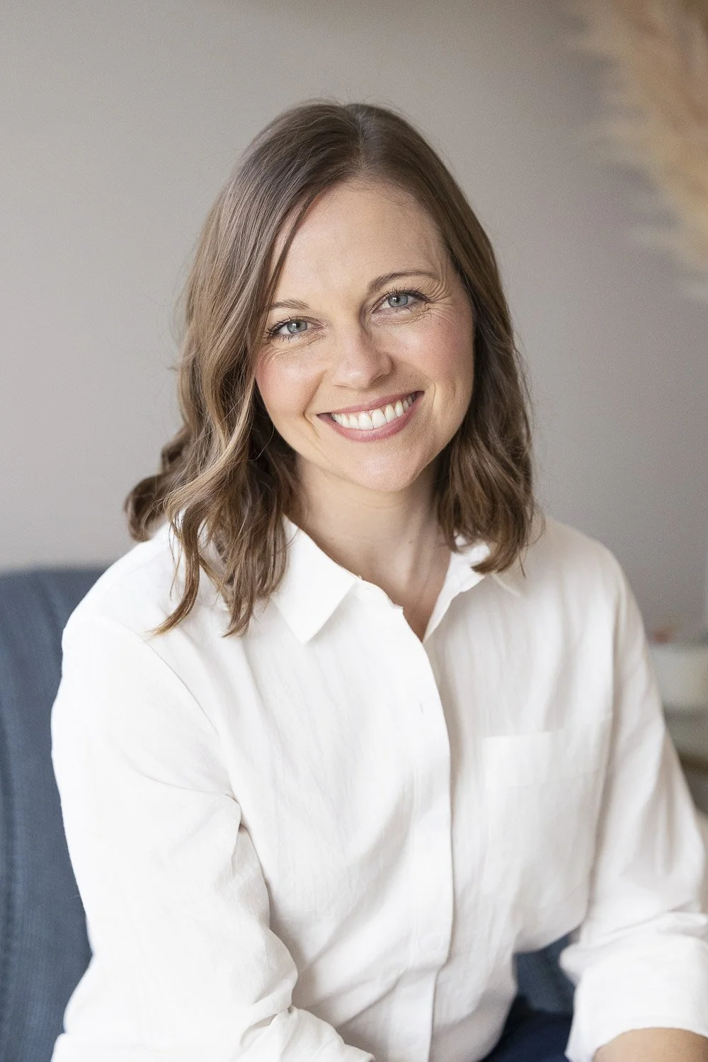 Portrait of a smiling woman with shoulder-length brown hair wearing a white shirt, sitting on a blue chair in a well-lit room.