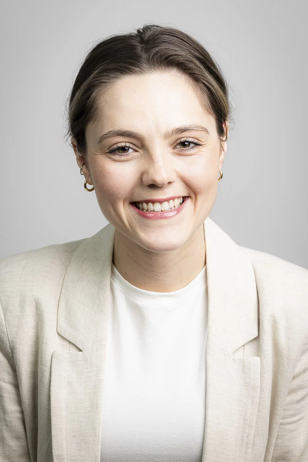 A smiling woman with short brown hair, wearing a cream blazer, against a light gray background.