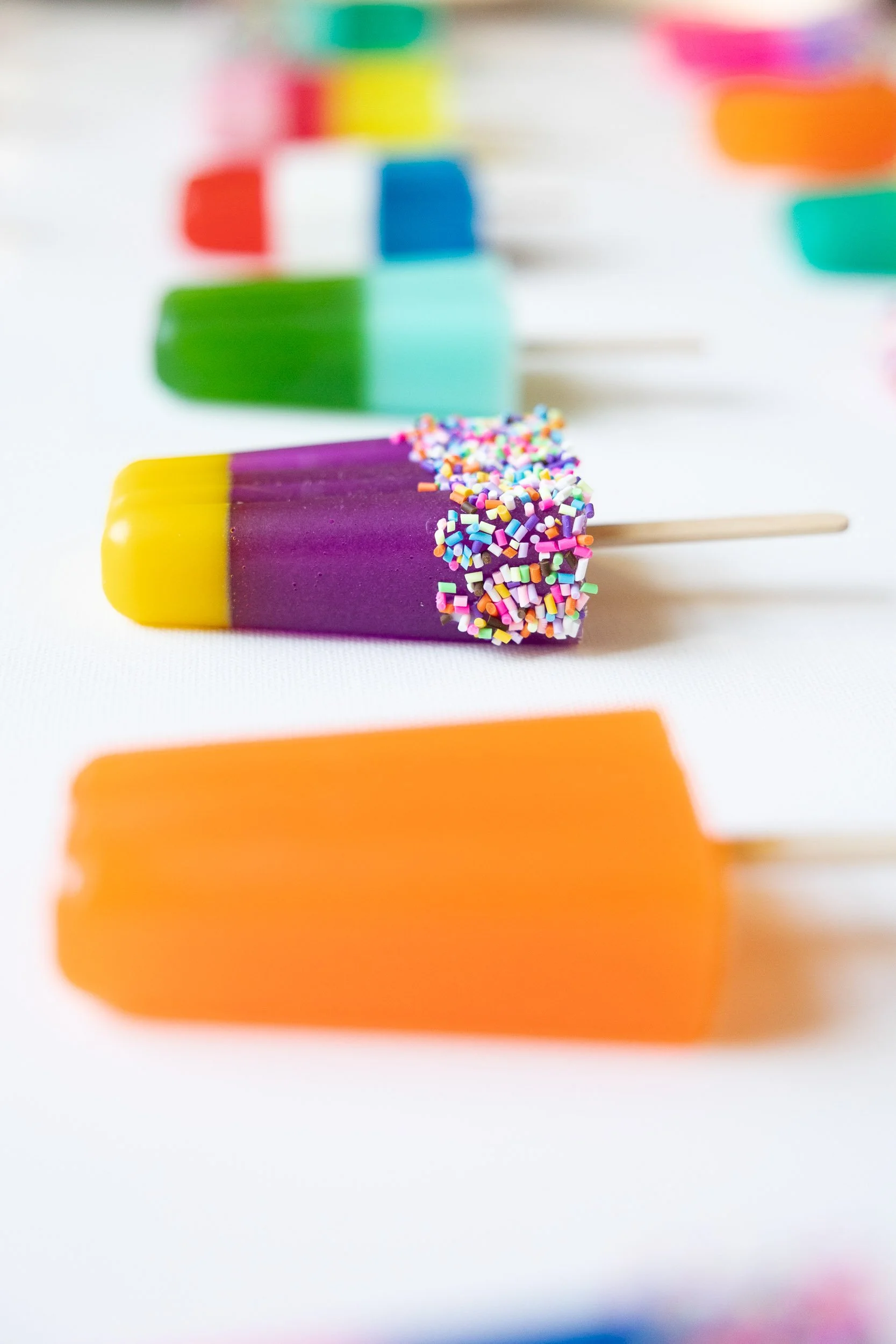 Colorful resin popsicles with vibrant colors including orange, purple, yellow, green, blue, and red, layered with sprinkles on a white surface.
