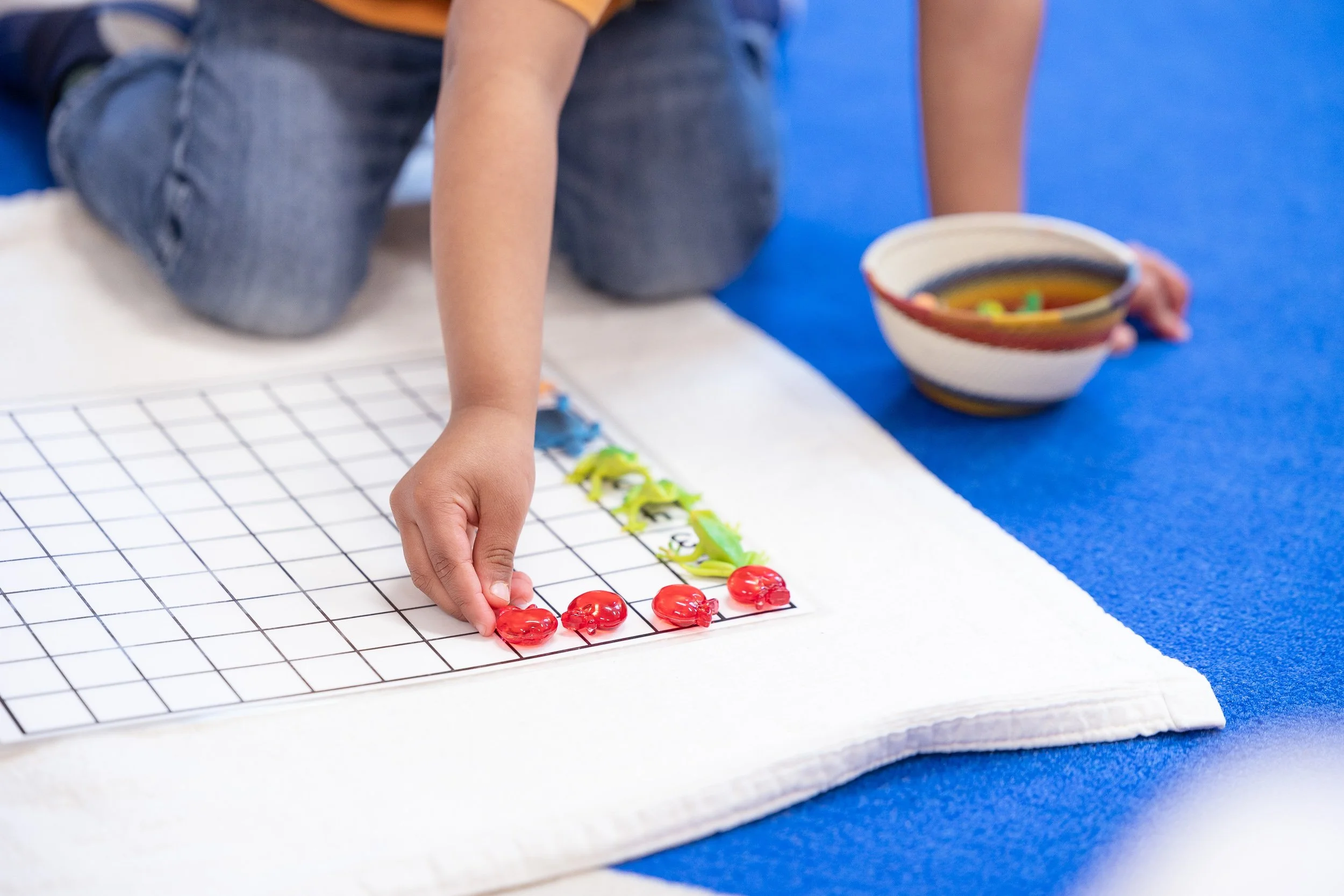 Child playing with colorful plastic toys on a grid board on a white cloth at a Montessori school in Boston.