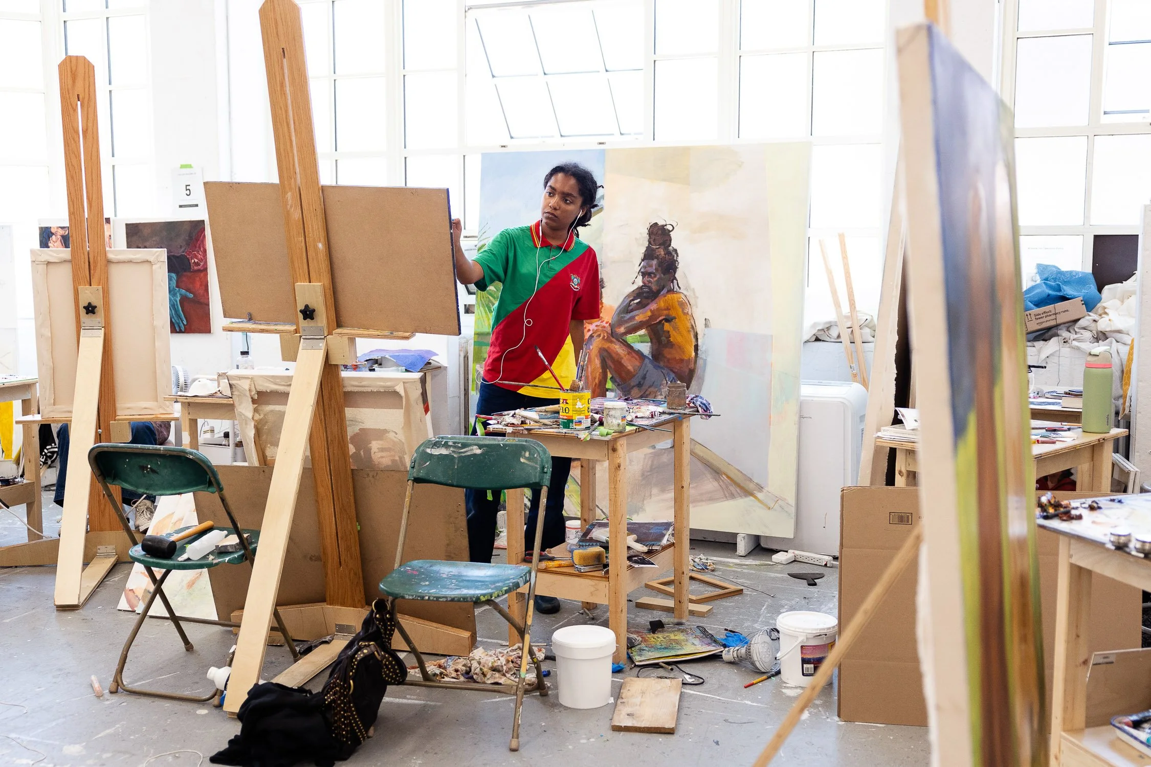 An artist in a studio painting on an art school campus in Providence, Rhode Island. The studio is cluttered with art supplies, easels, chairs, and partially finished artworks.