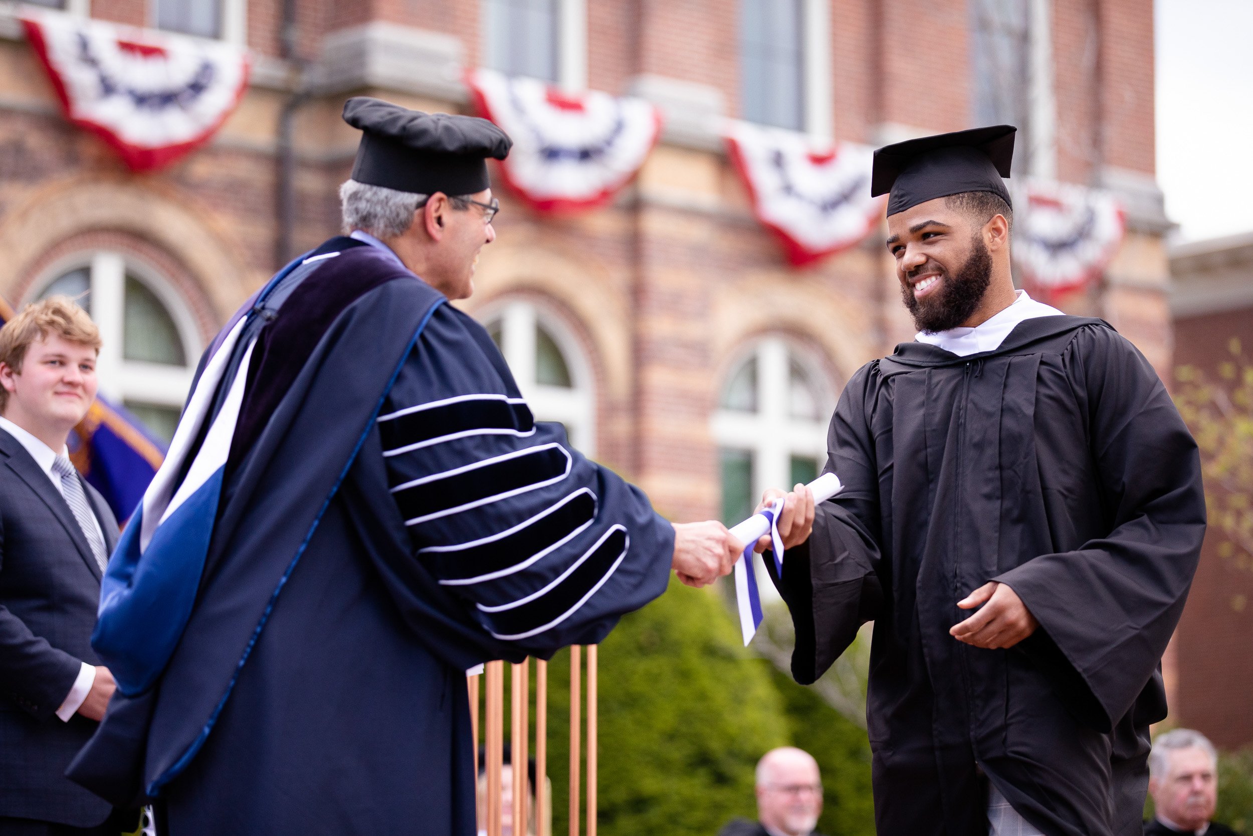 A graduate in cap and gown receiving his diploma from college president in outdoor commencement ceremony.