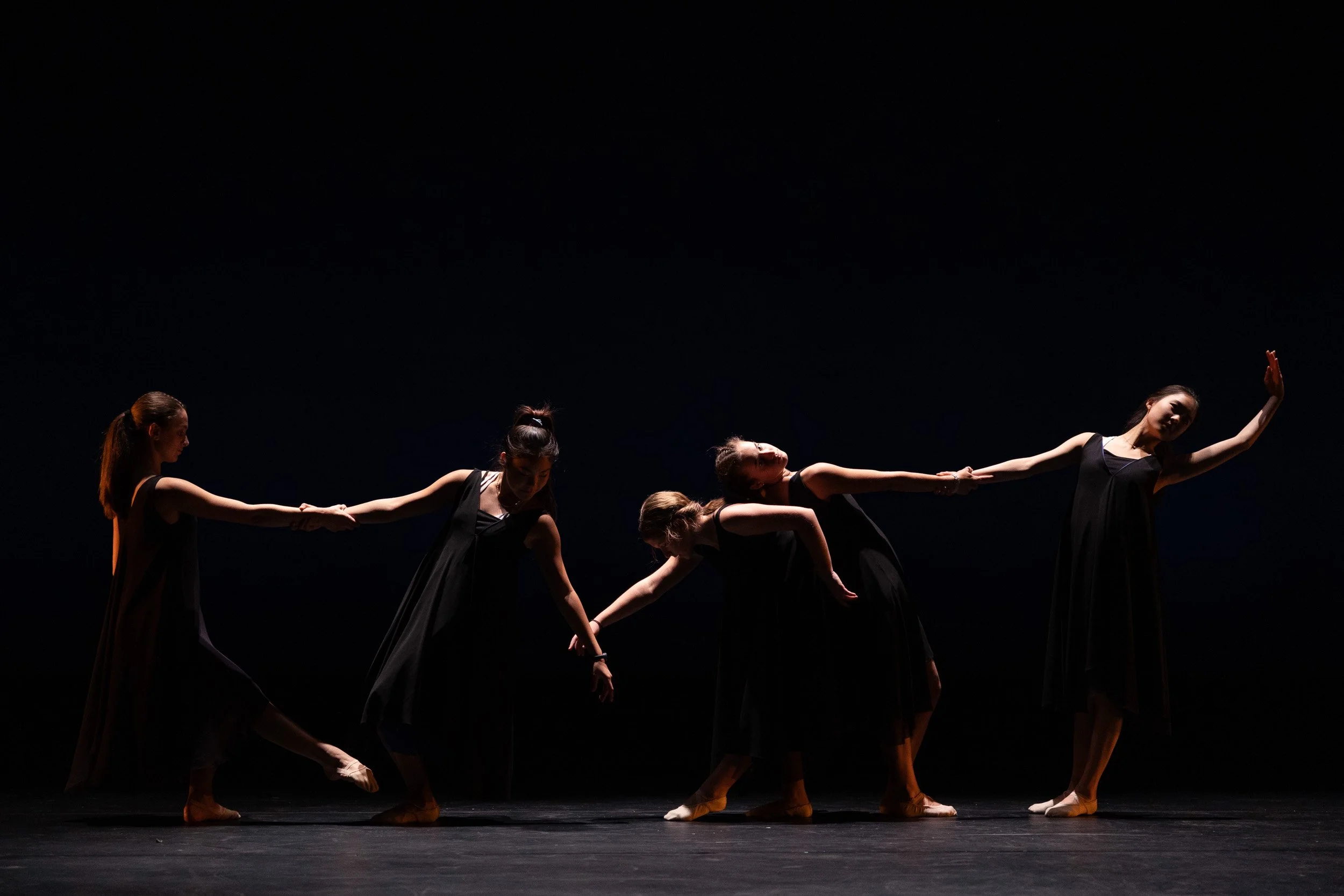 Five female ballet dancers in black dresses holding hands on stage with dark background during a dance recital in Boston, Massachusetts.