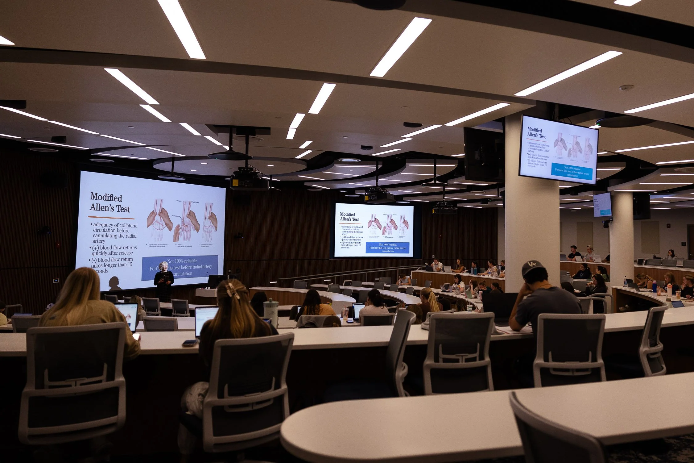 A large college lecture hall with multiple rows of curved desks filled with students and attendees, facing a stage with a presenter and multiple large screens at a university in Massachusetts.