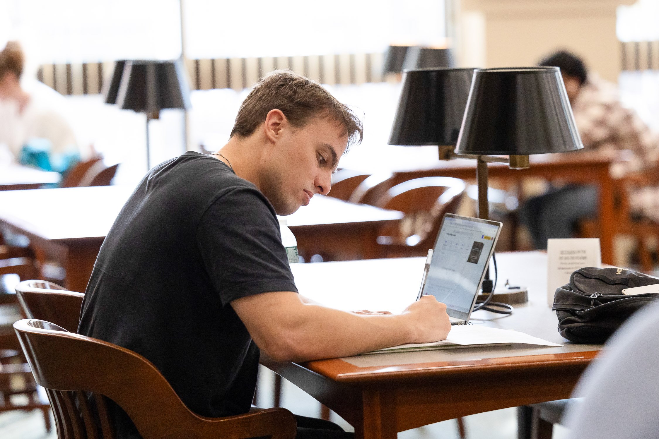 A young man sitting at a wooden table in a library or study area, working on a laptop with a notebook and pen in front of him. There are multiple lamps and chairs in the background.