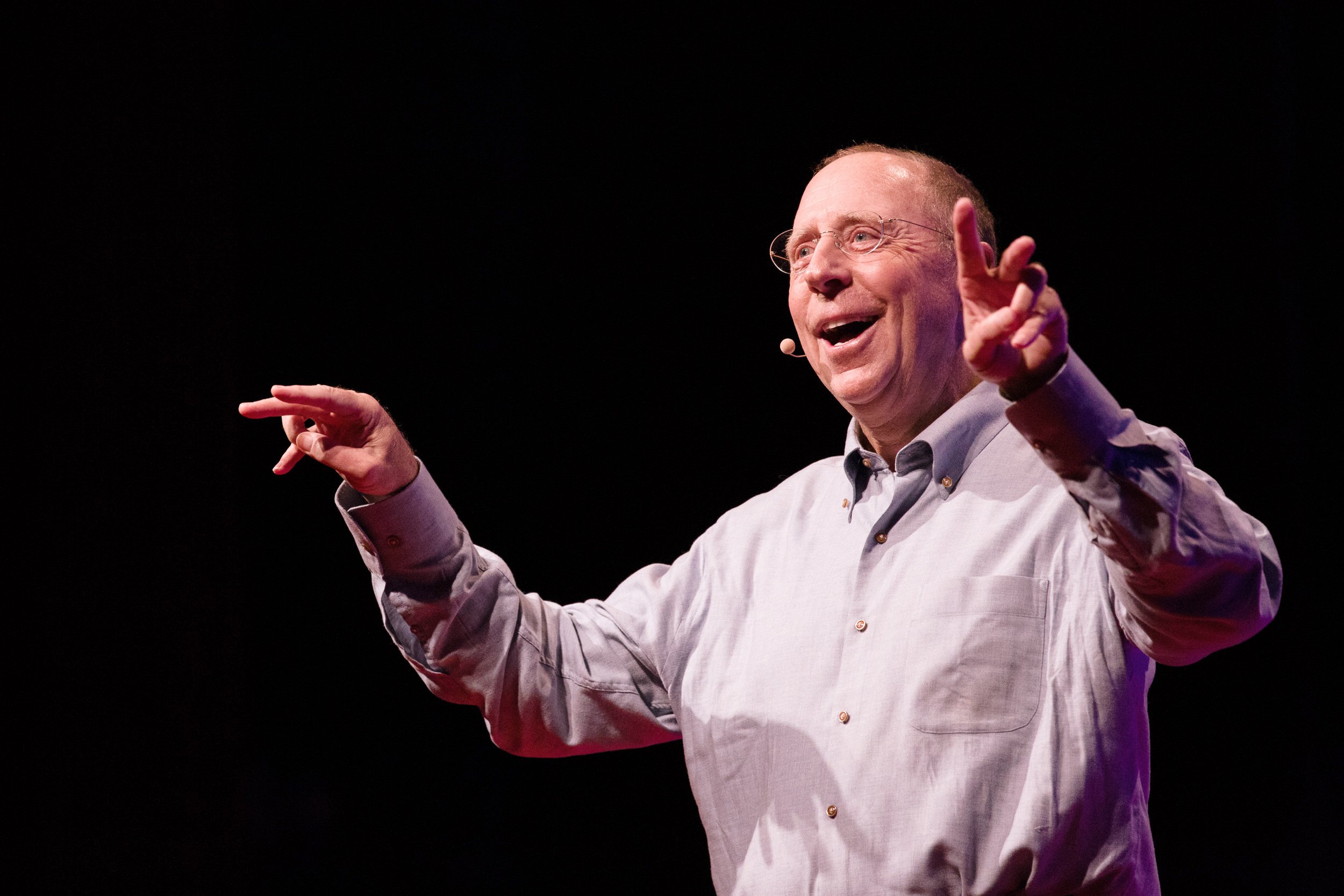 Man speaks on stage, gesturing with both hands, smiling, during Boston TEDx talk.