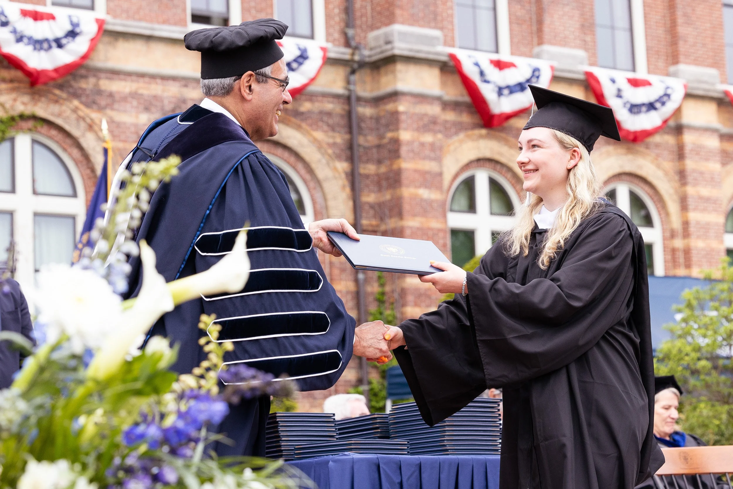 A young woman in a graduation gown and cap shaking hands with a man in academic regalia during a graduation ceremony outdoors on a Boston college campus.