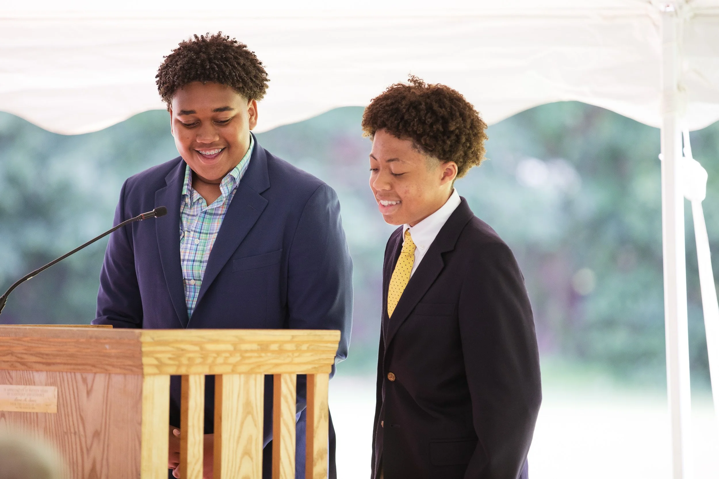Two middle school boys in suits standing at a wooden podium and smiling during Charles River School graduation ceremony.