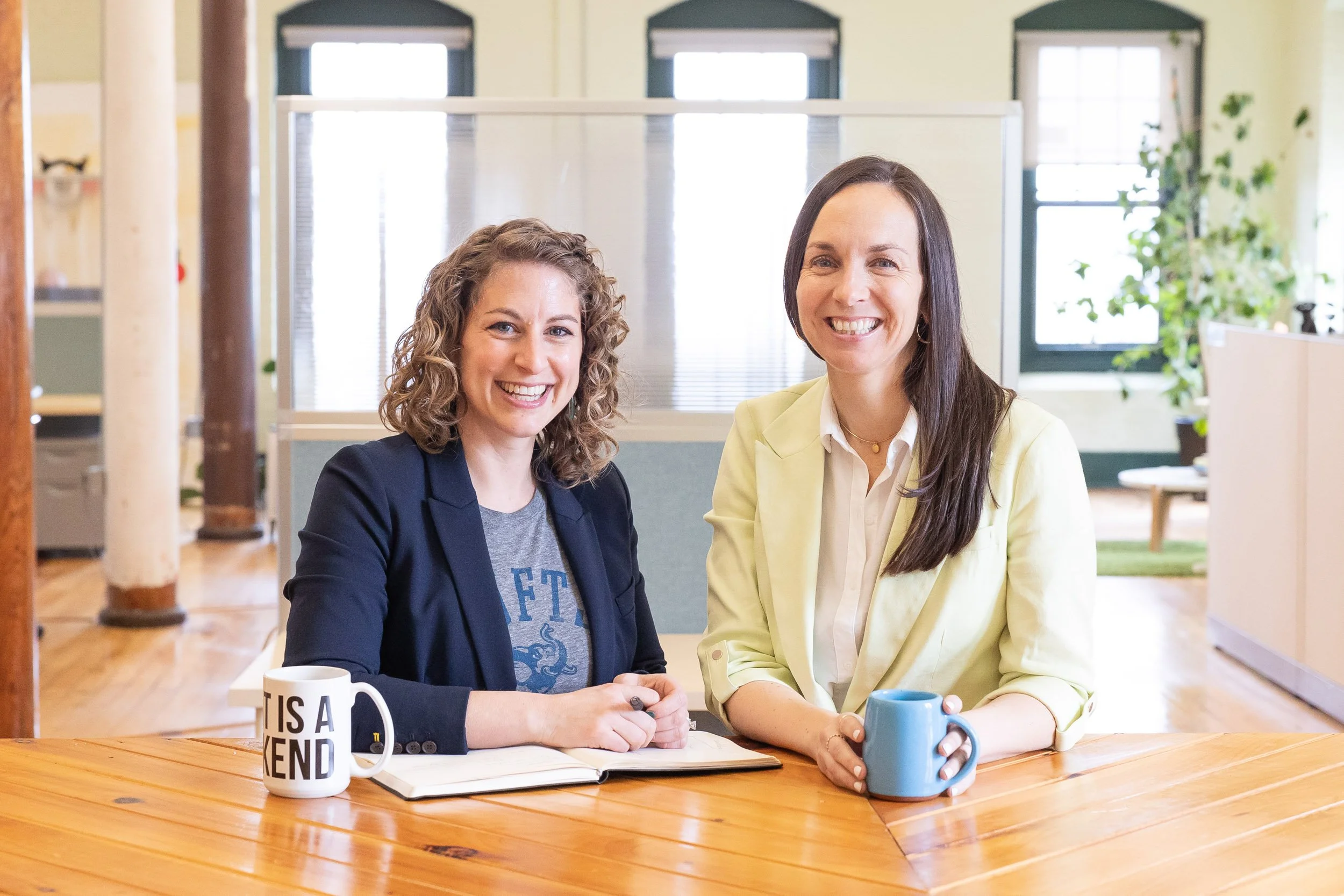 Two female business owners smiling and sitting at a wooden table in a bright, open Massachusetts office space.