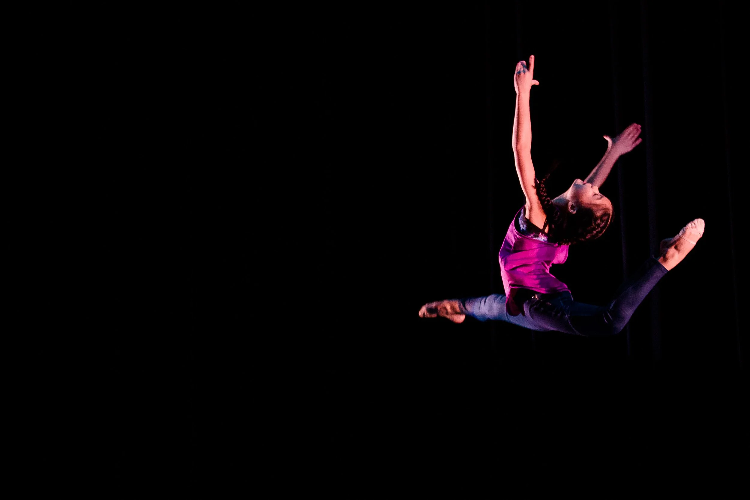 A dancer in a pink top performs a leap against a black background at a Massachusetts high school dance recital.