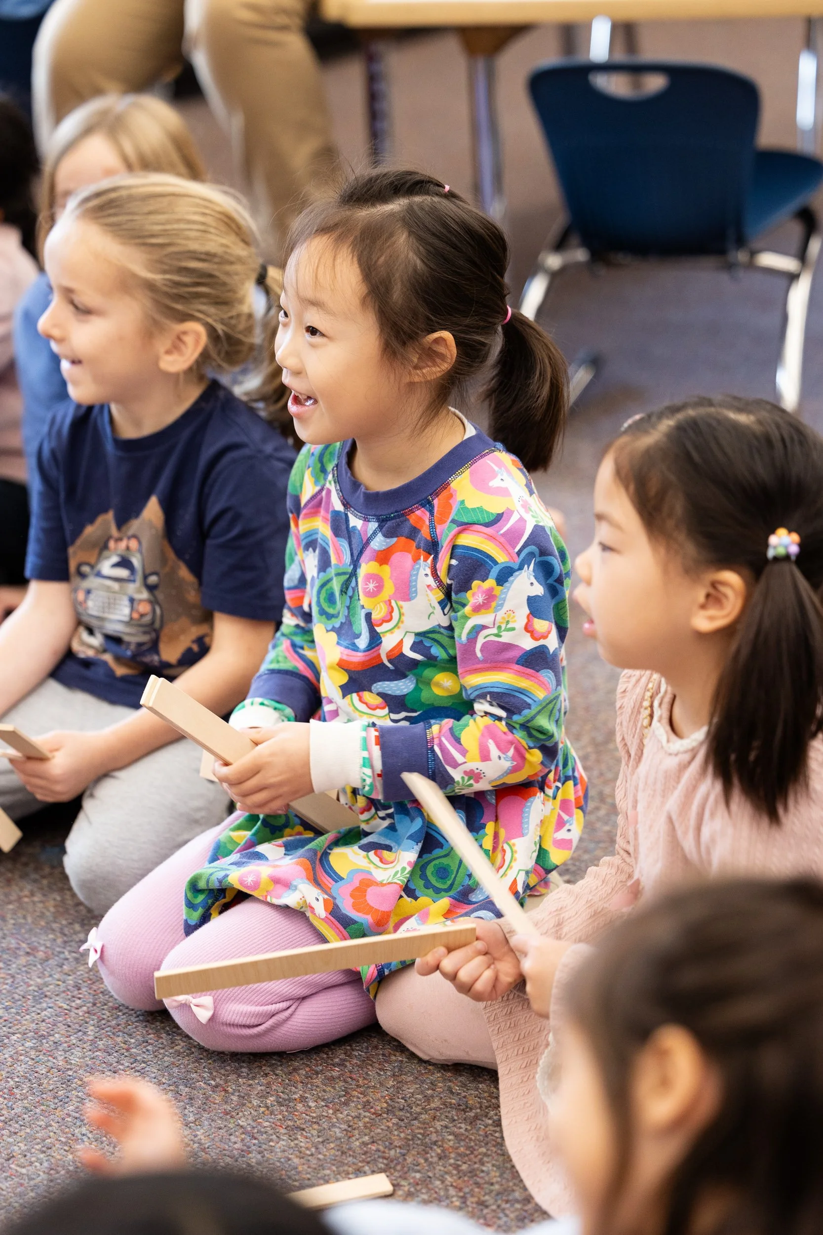 Elementary school children sitting on the floor, smiling and holding drum sticks during music class.