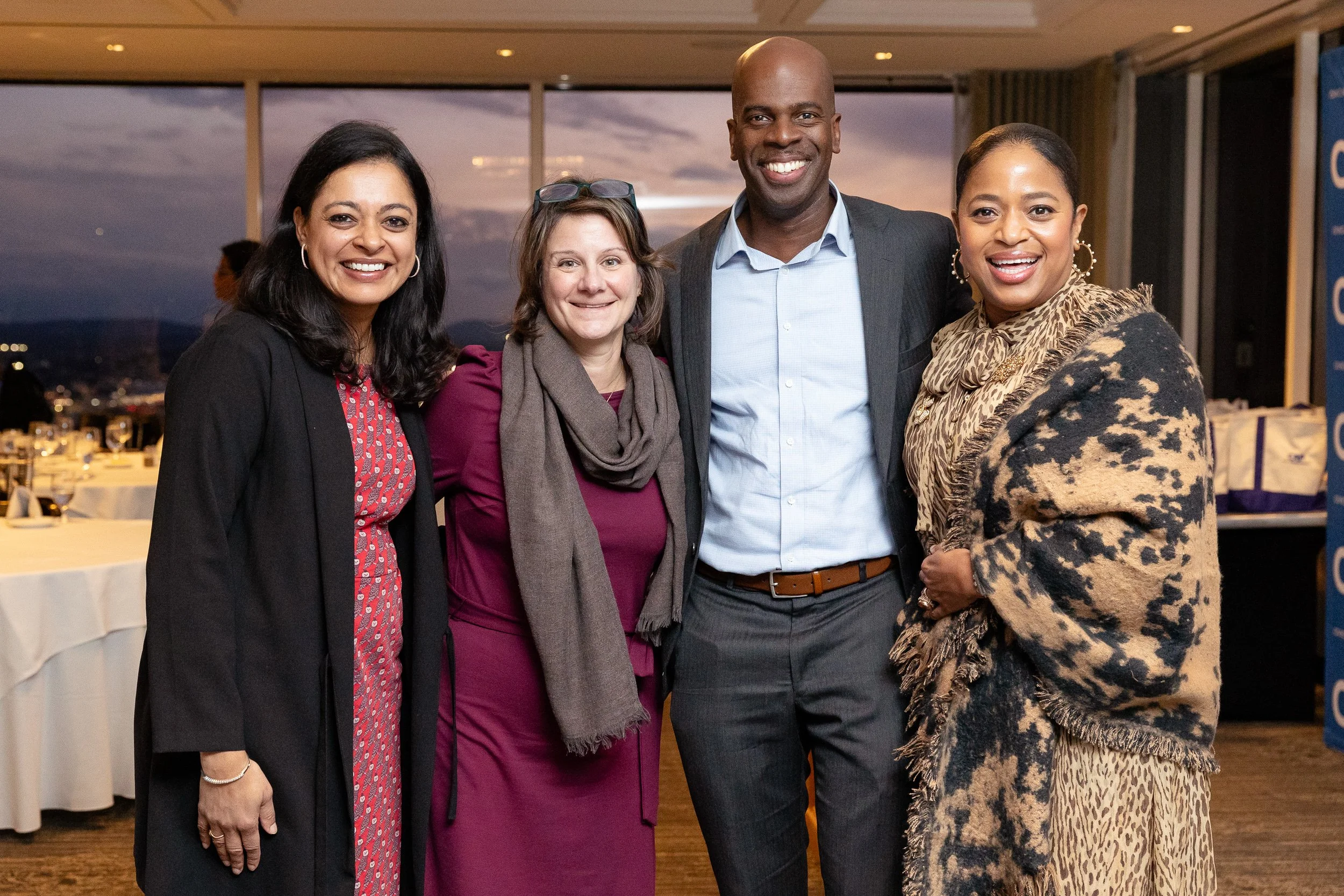 Group of people smiling and posing for a photo at an indoor event with a Boston cityscape view through large windows in the background.