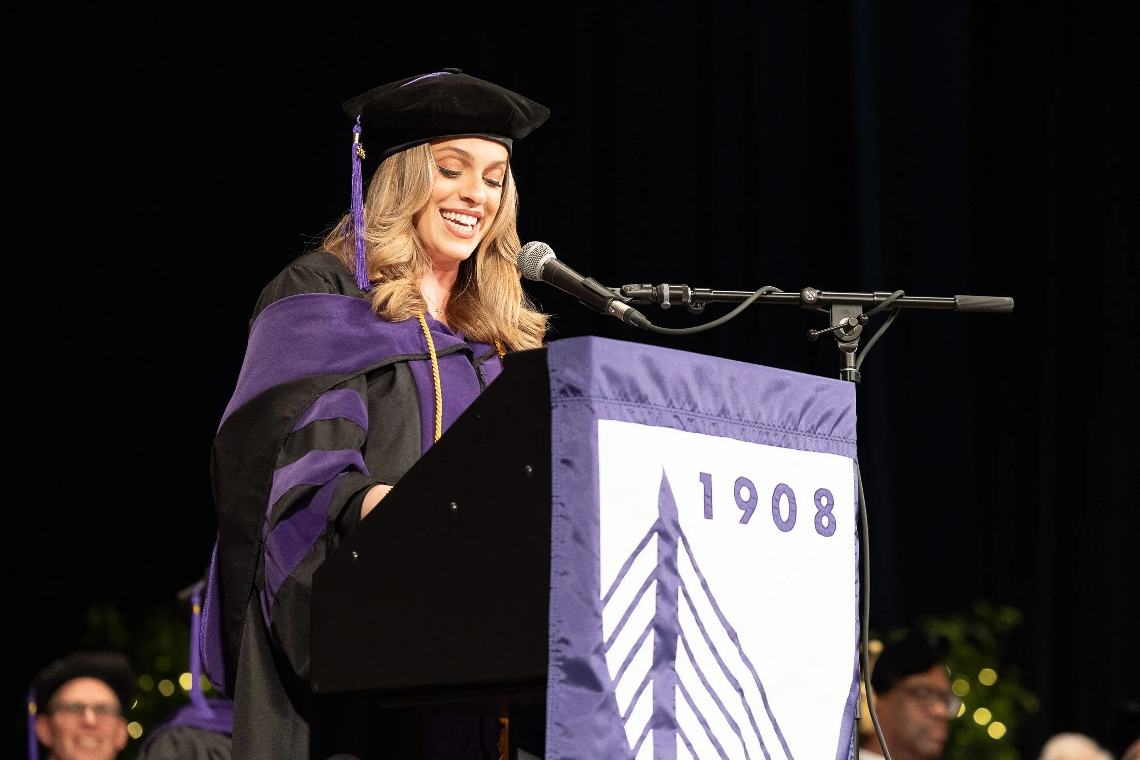 A woman in cap and gown speaking at a graduation ceremony behind a lectern at the Wang Theater in Boston.