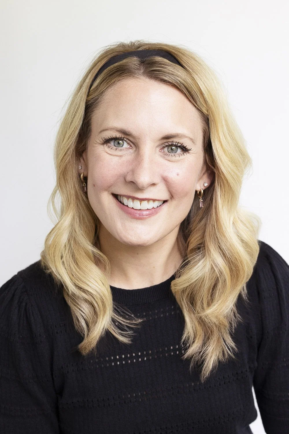 A woman with blonde hair, wearing a black headband and shirt, smiling against a plain white background.