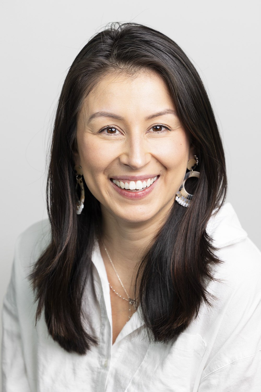 Headshot of a woman with straight, dark hair, smiling, wearing a white shirt against a plain light gray background.