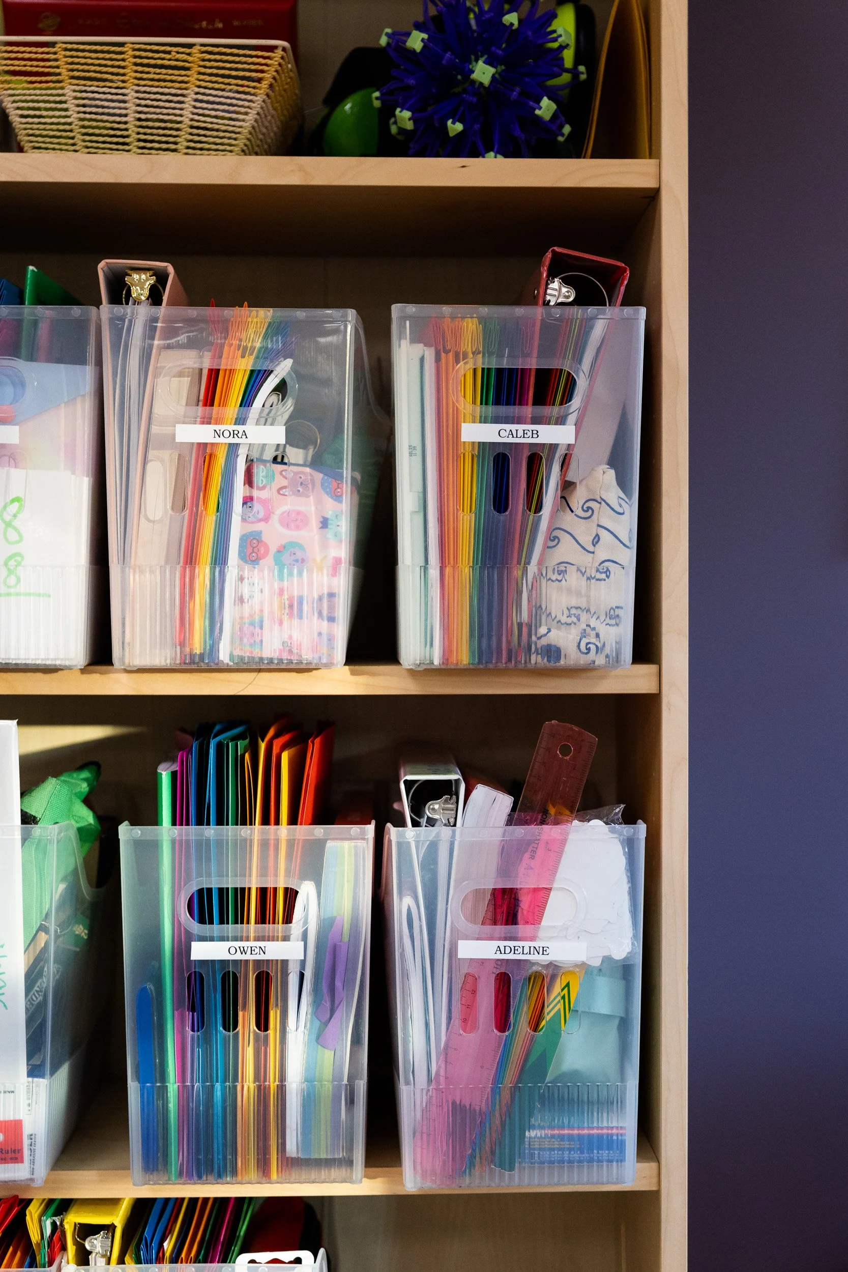 Clear plastic bins with colored pencils, markers, and writing supplies on a wooden shelf in a Montessori classroom.