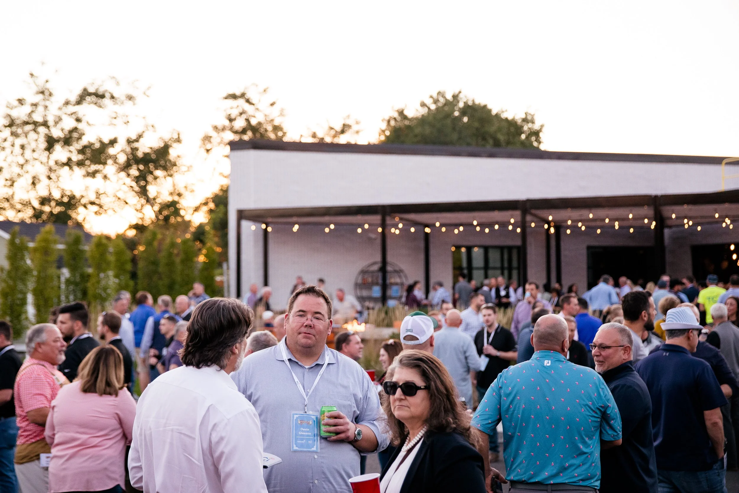 People gathered outdoors at a company party during sunset, with string lights hanging from a modern building.