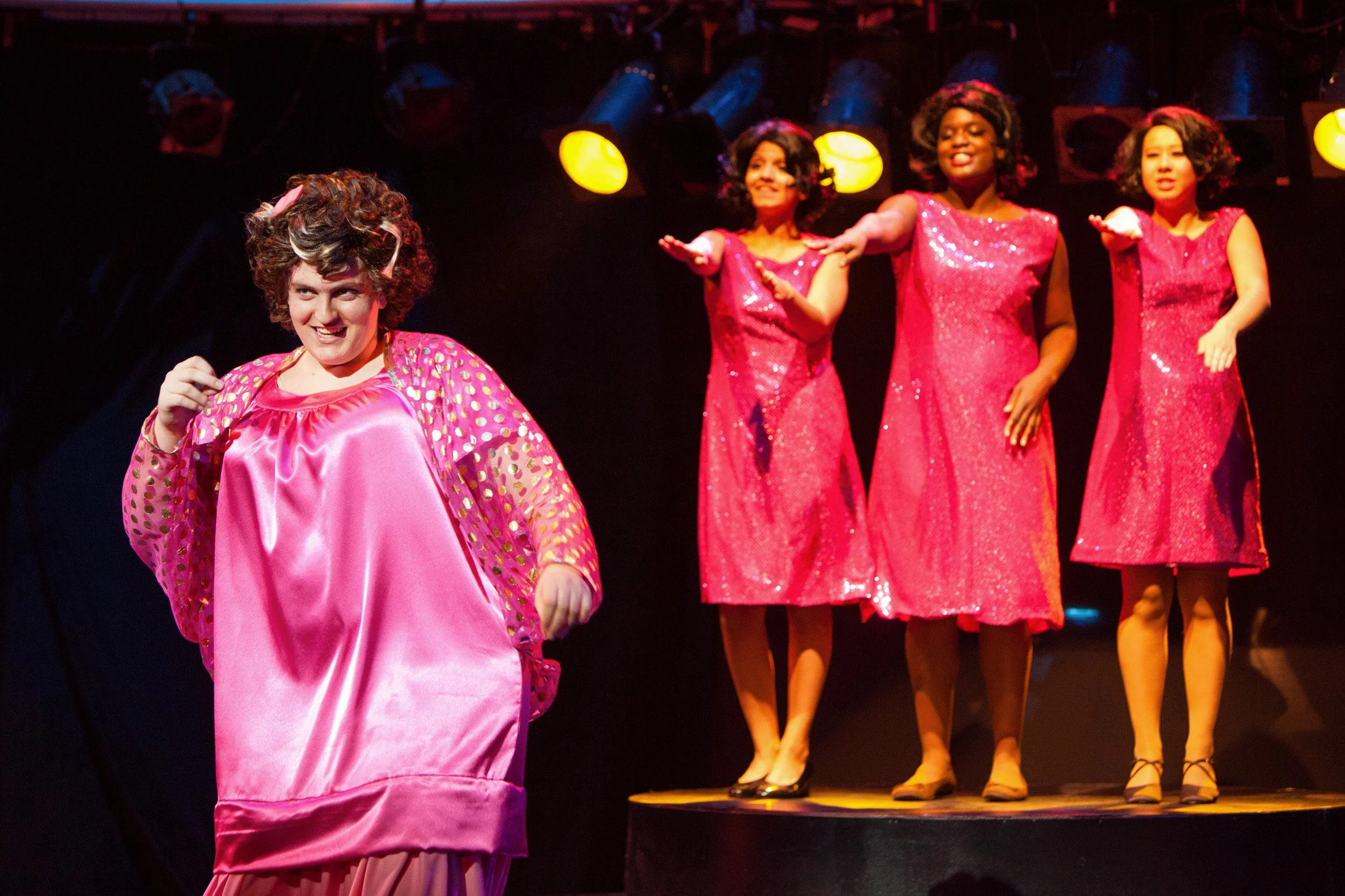 Four women dressed in pink costumes performing on stage in a high school production of Hairspray.