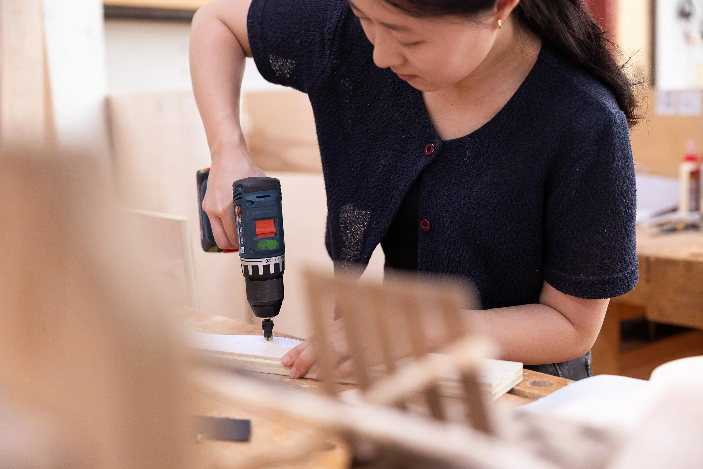 An art student using a cordless drill to assemble a piece of furniture in a woodworking workshop.