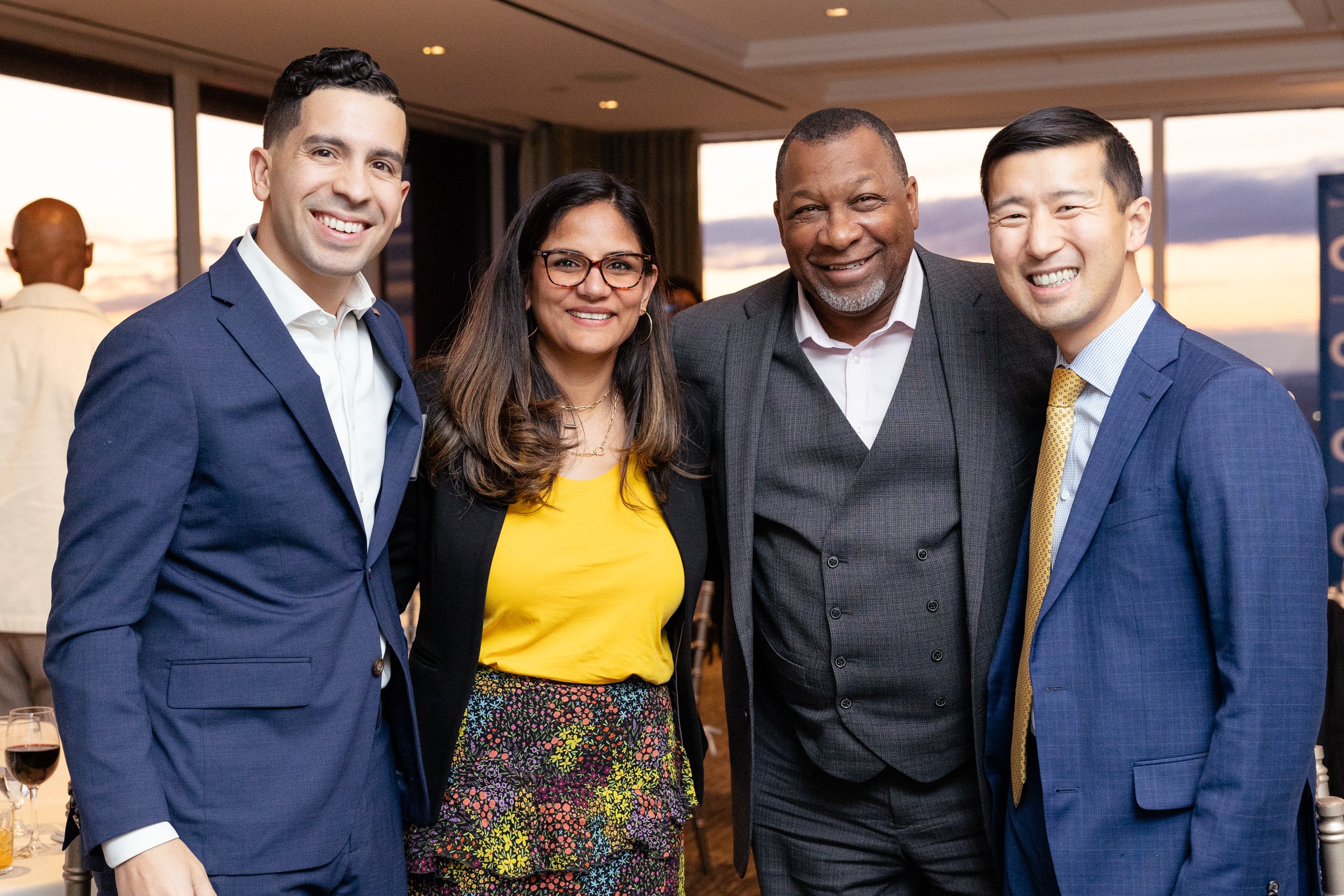 Five professionals dressed in business attire, smiling at a social or networking event during sunset.