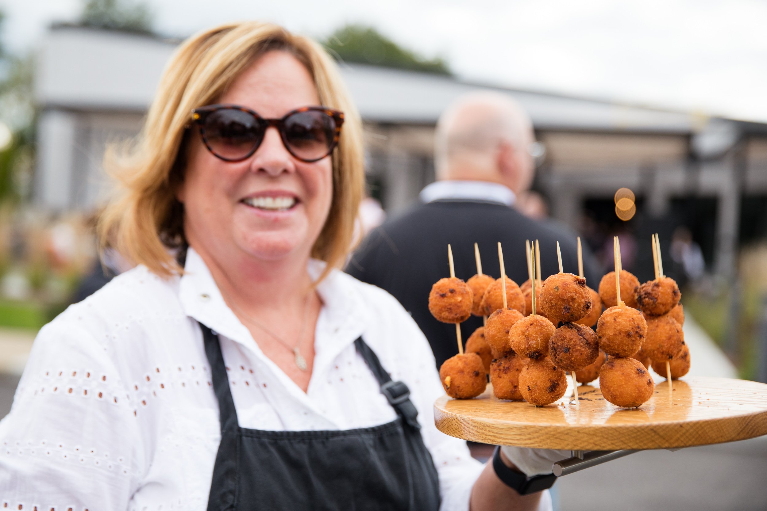 Woman wearing sunglasses and an apron holding a wooden tray with fried food at an Oktoberfest event.