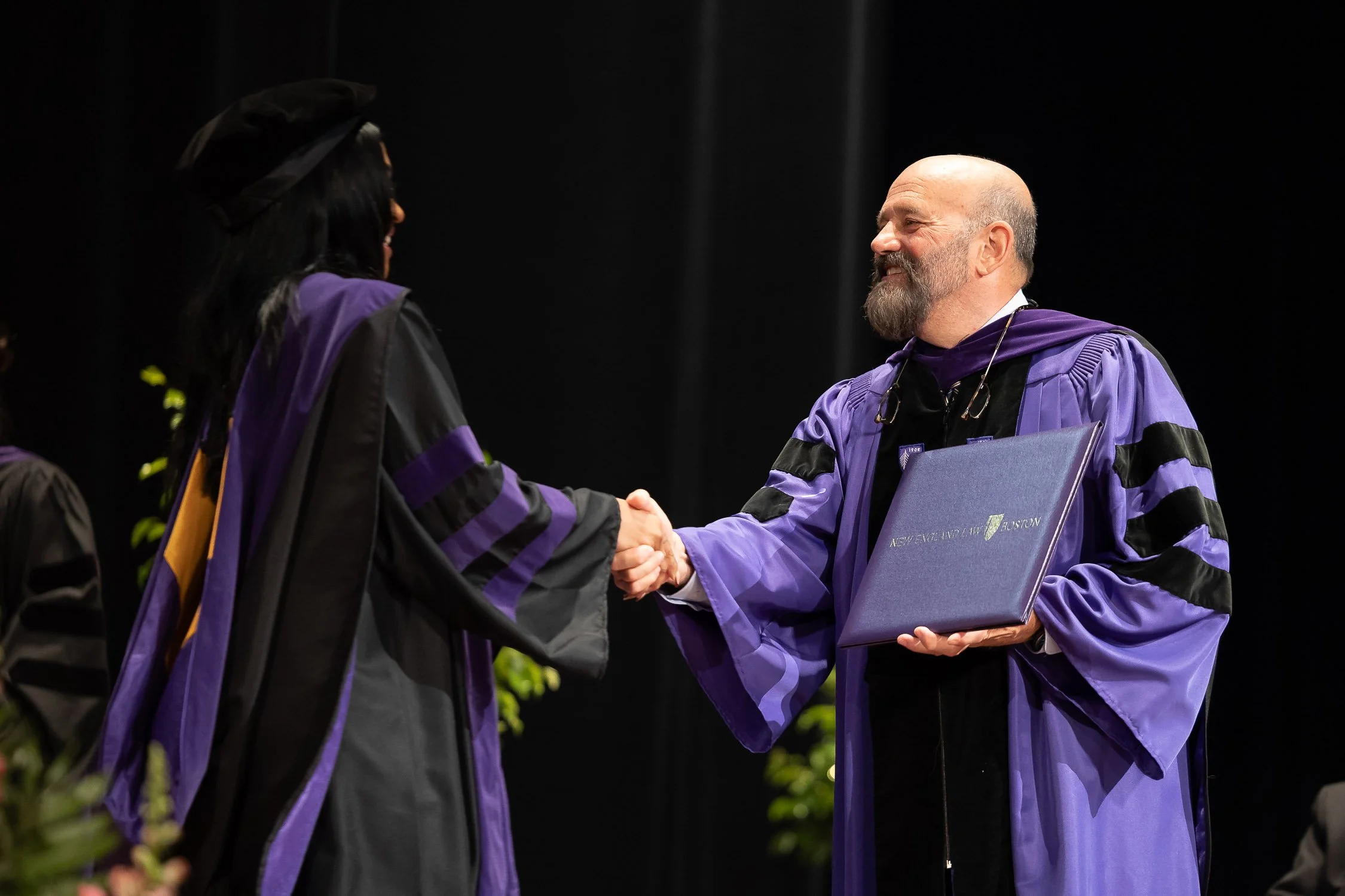 A graduate shaking hands with a man in a purple gown holding a diploma during a graduation ceremony.