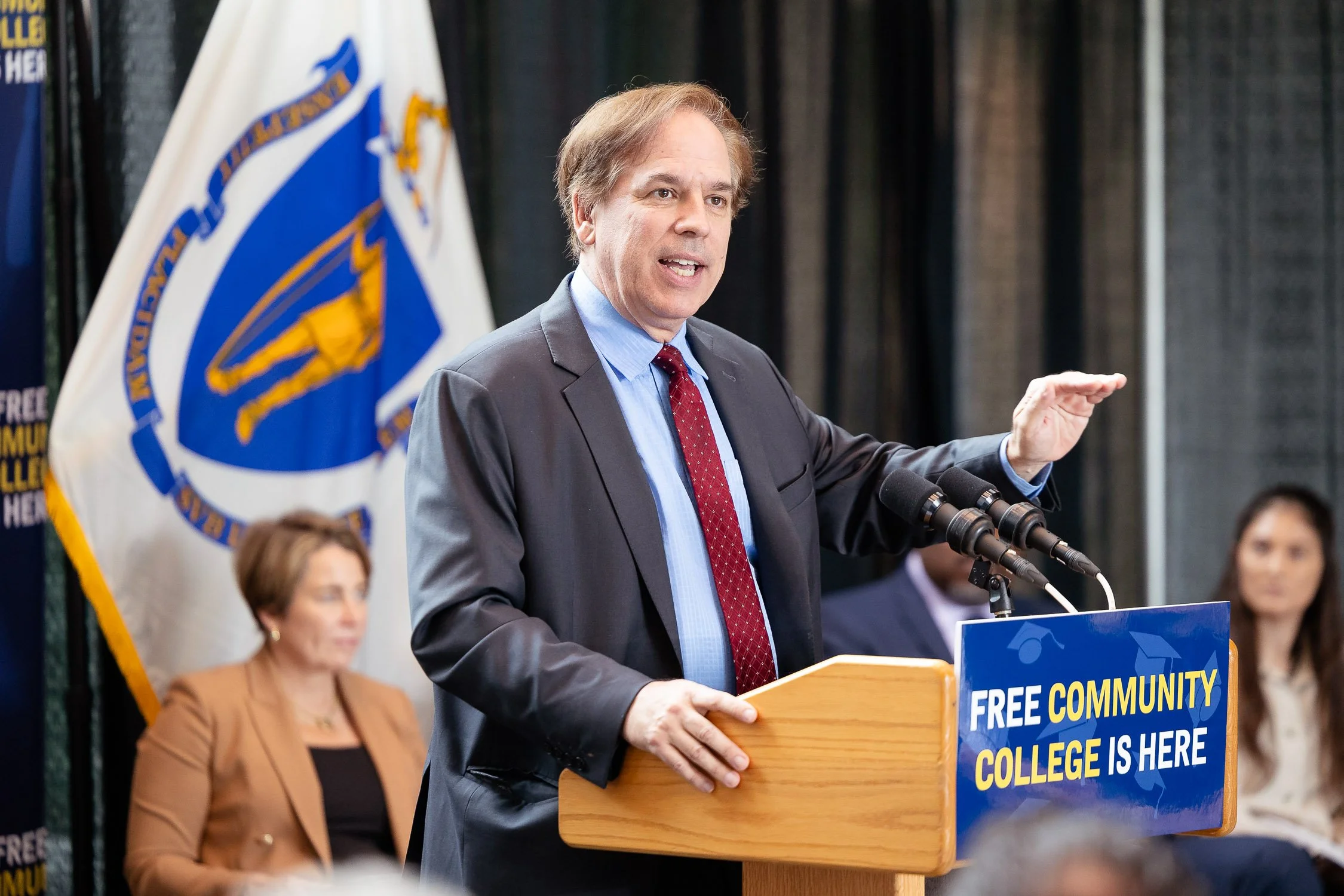 Man in suit and red tie speaking at a podium with a sign reading 'Free Community College Is Here' at MassBay Community College in Framingham.