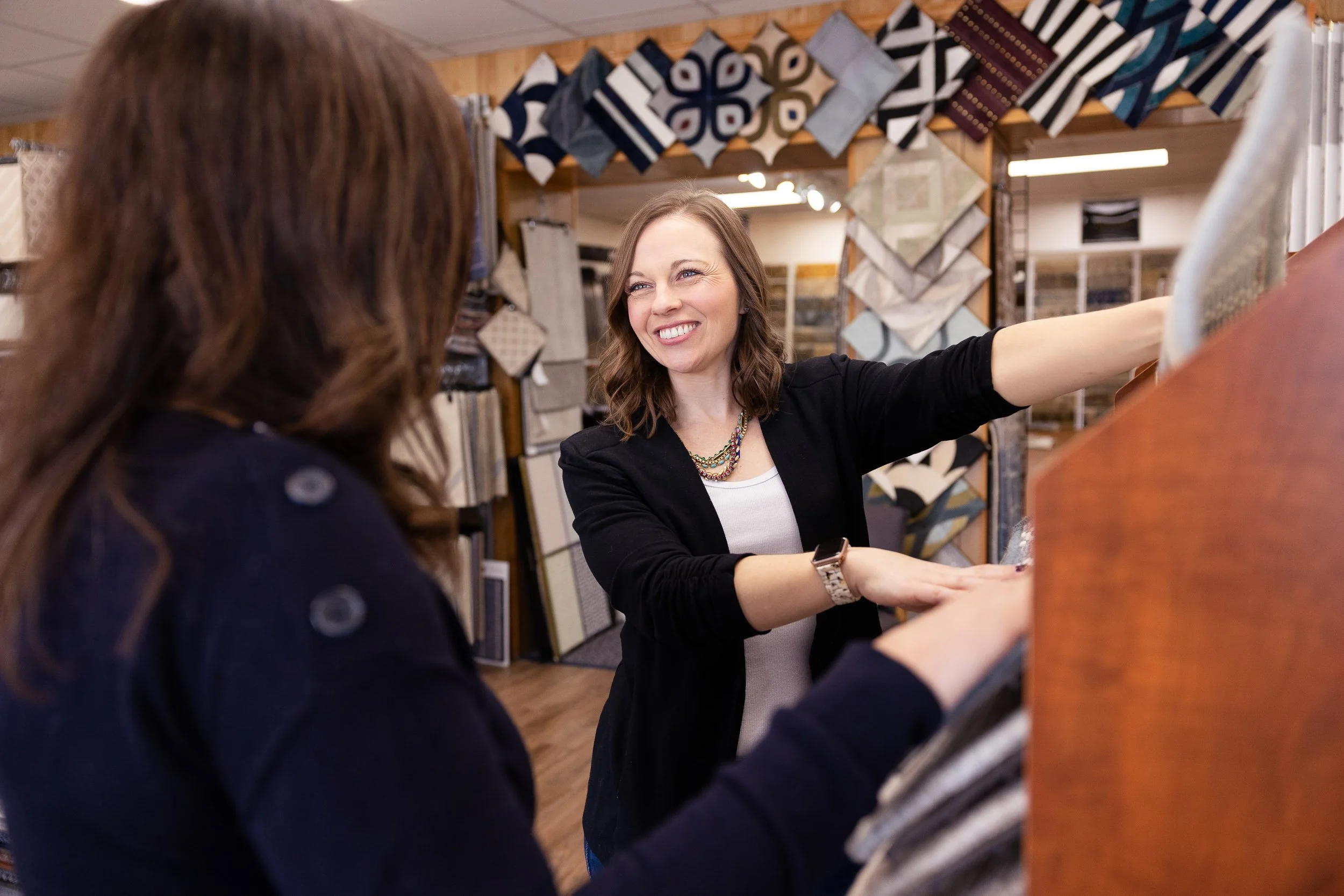 A female interior designer smiling and showing a fabric sample to another woman in a home decor store in Needham, Massachusetts.