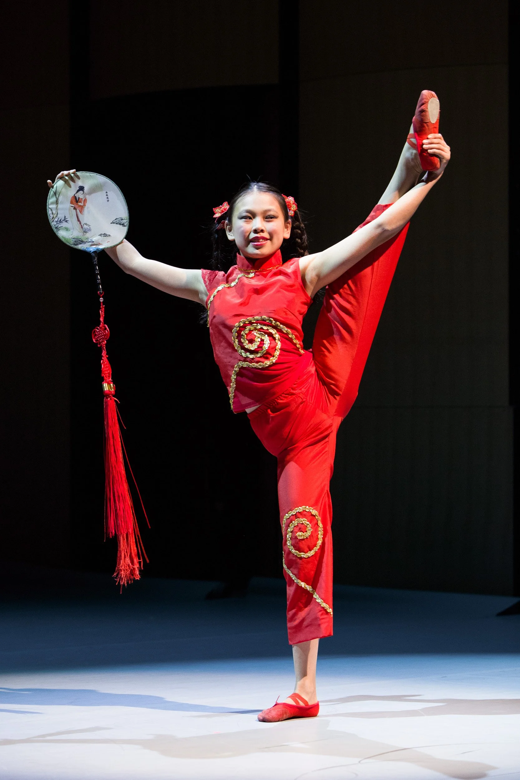A young girl in a red traditional Chinese costume performing a dance on stage at a high school dance performance in Massachusetts.