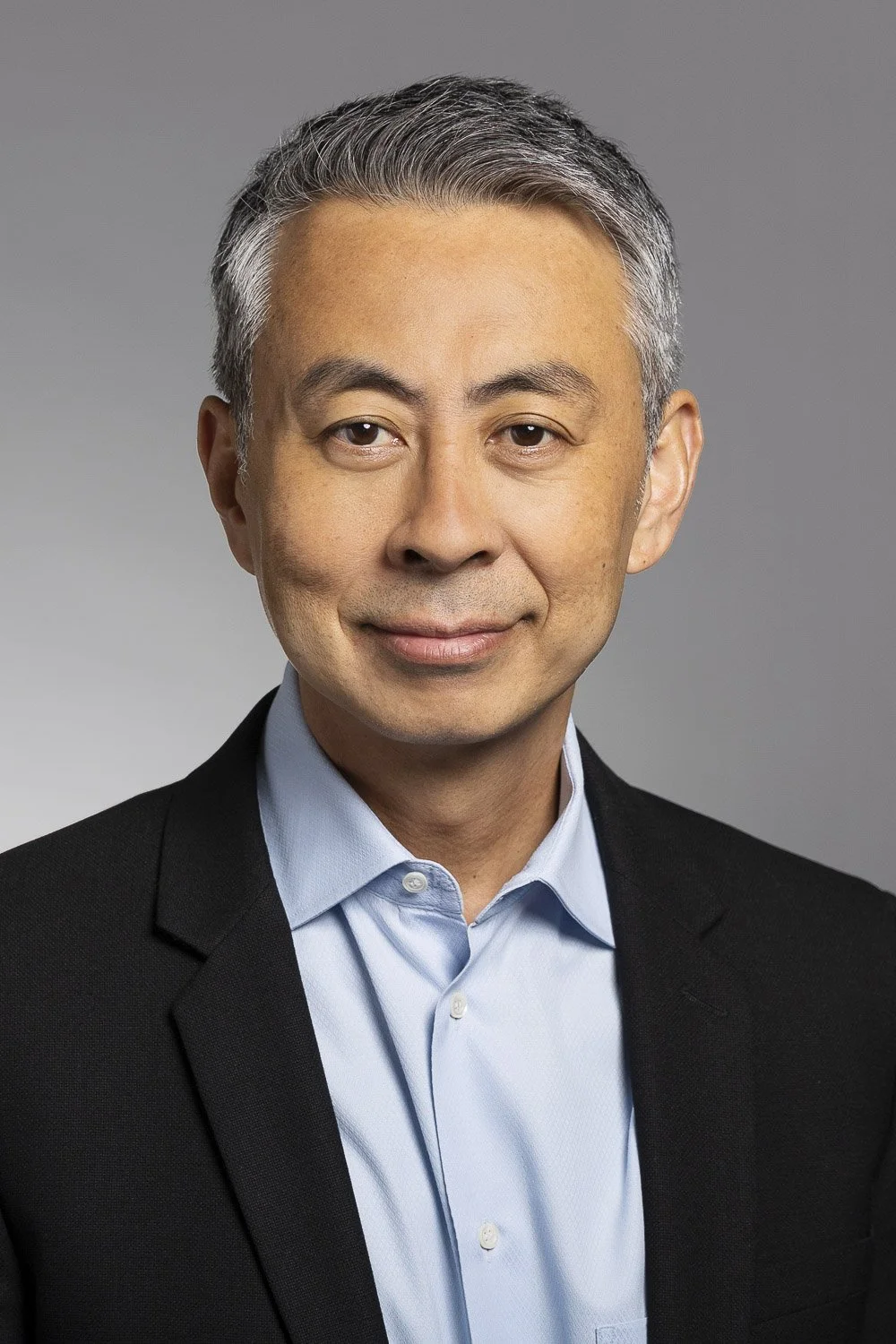 Headshot of a biotech executive with short gray hair, wearing a light blue dress shirt and dark blazer, against a gray background.