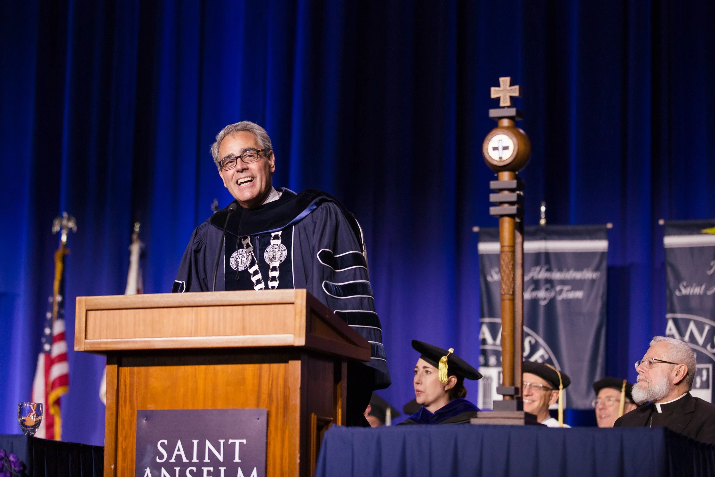 A man in academic regalia speaking at a podium during a Saint Anselm College event in New Hampshire.