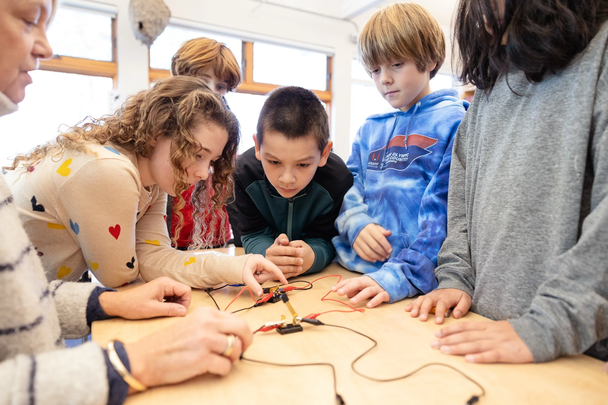 A group of students gathered around a table, focused on an electronics project involving wires and small components at a private Boston middle school.