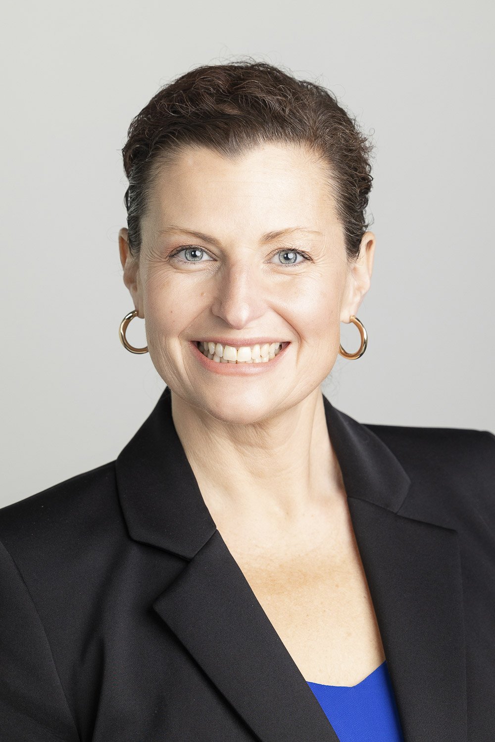 A professional headshot of a woman with short brown hair, wearing a black blazer and a blue top, smiling at the camera against a plain light background.