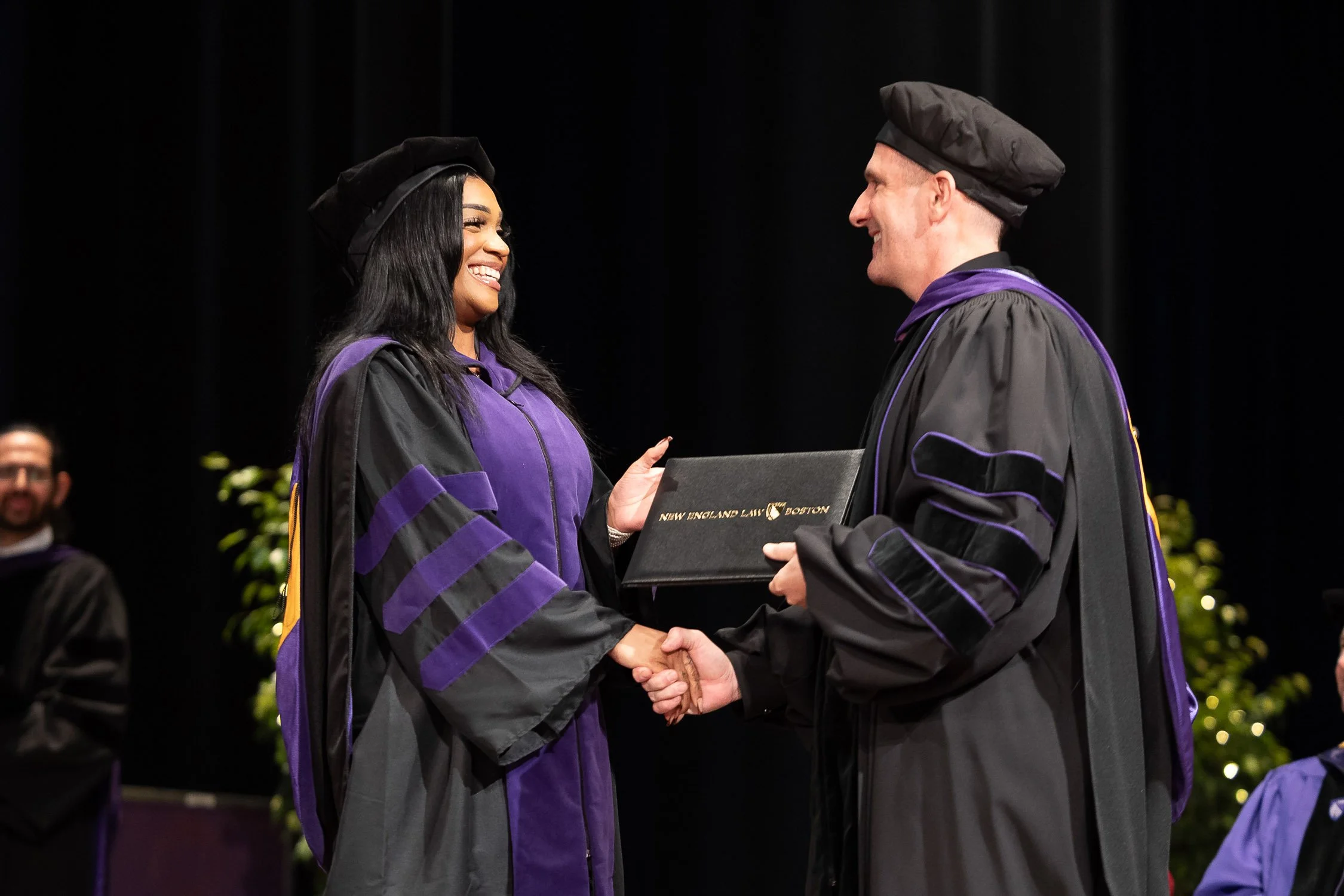 A woman in academic regalia receiving a diploma during a graduation ceremony in Boston.