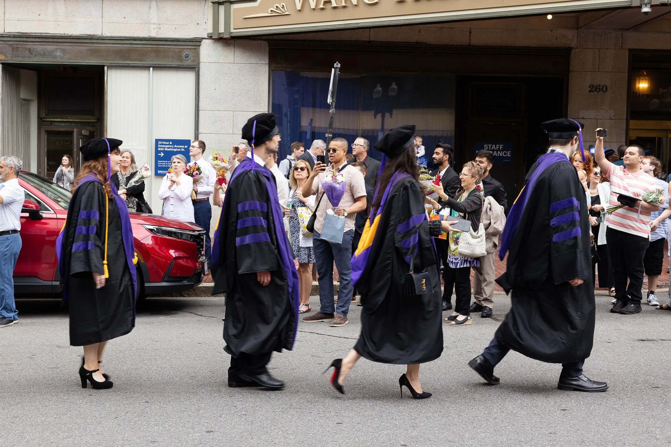 Group of graduates in black gowns walking in a procession on a Boston city street, with people watching and taking pictures on the sidewalk.