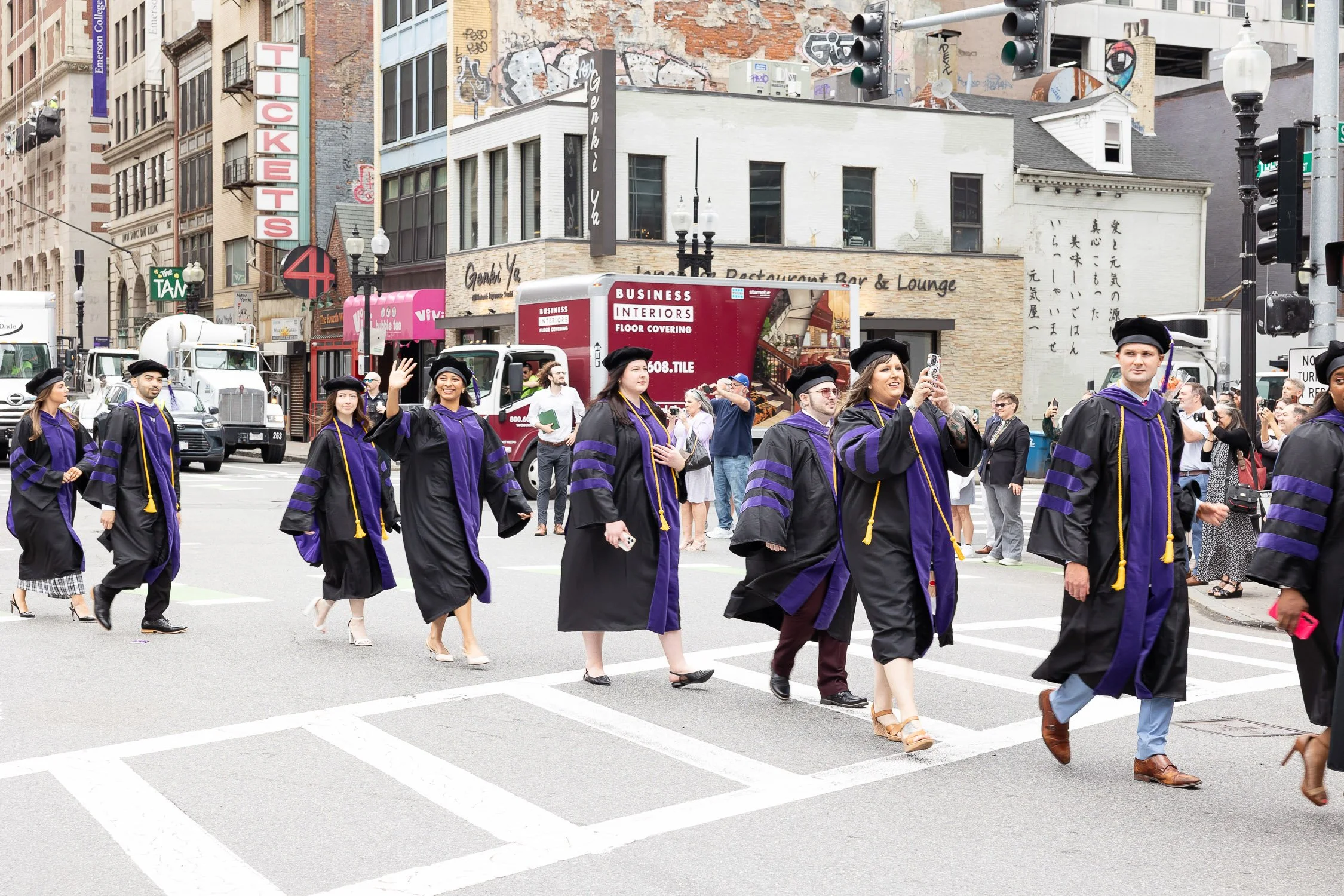 A group of graduates in academic regalia walking across a Boston city crosswalk during graduation procession.