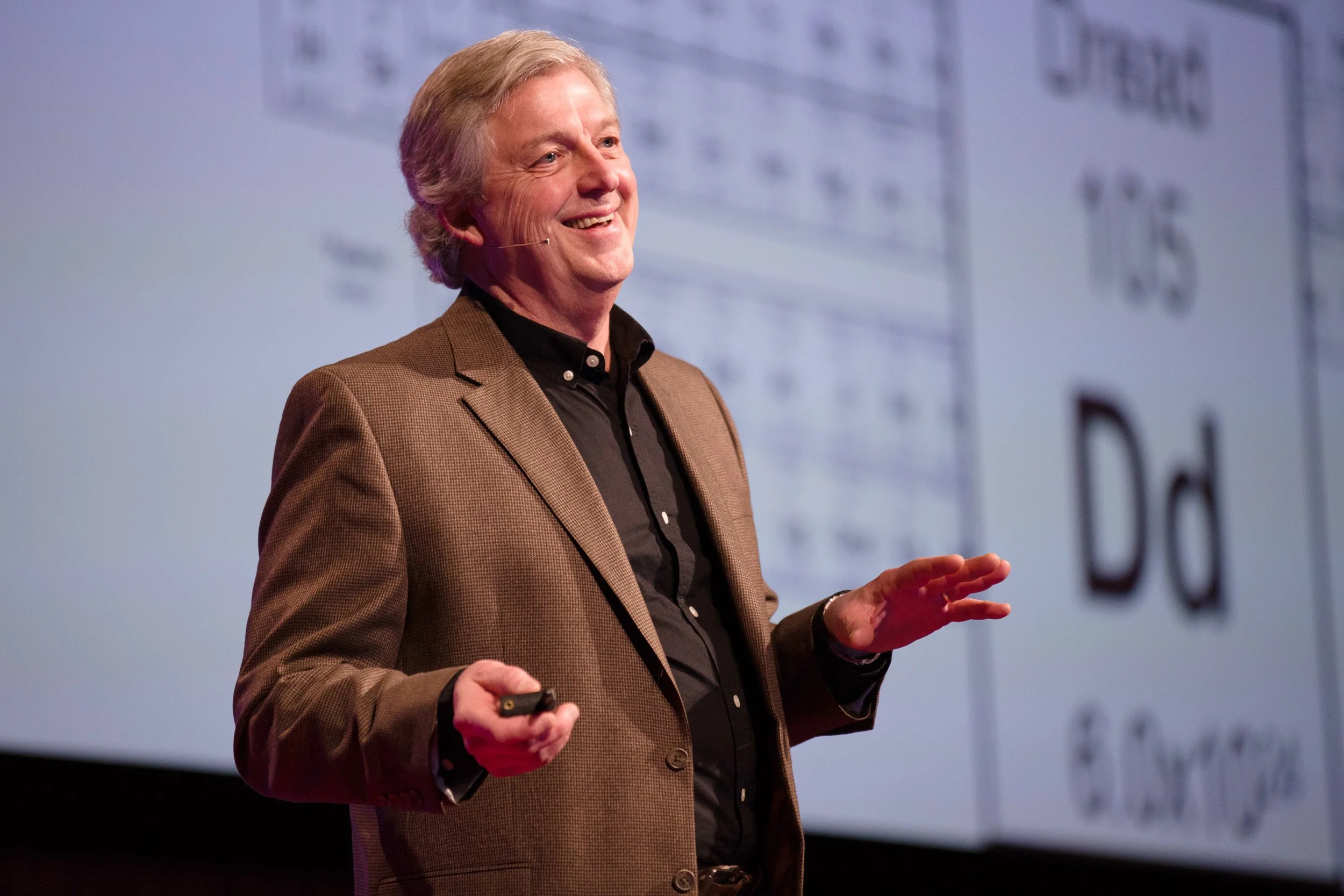 A man smiling and speaking in front of a large screen displaying a periodic table of elements during TEDx talk in Boston.