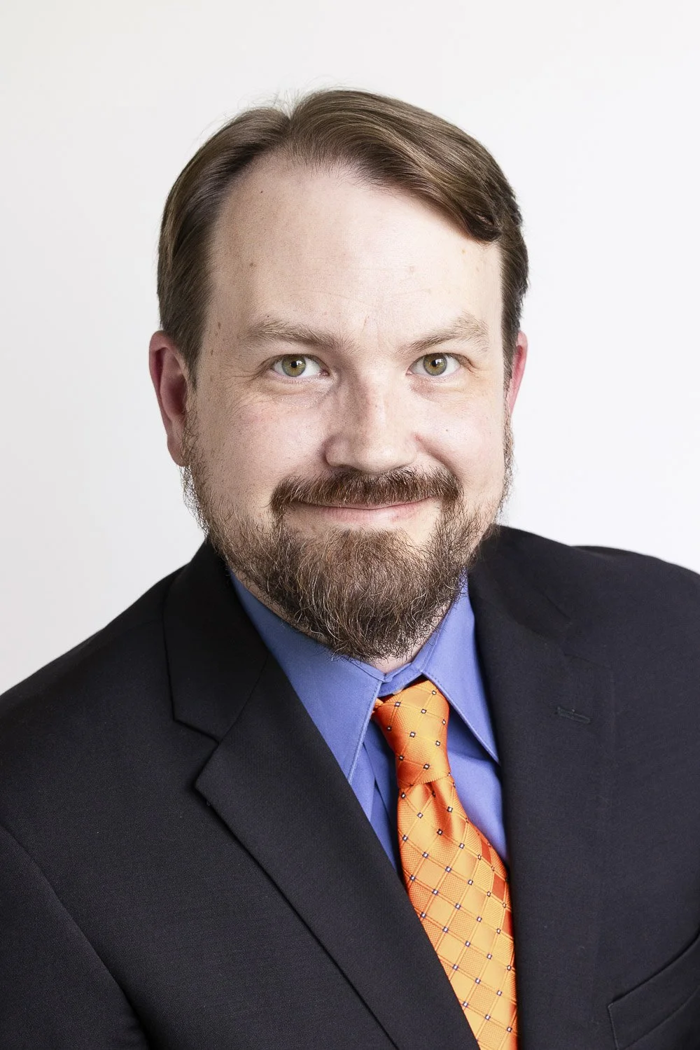 Headshot of a man with a beard, mustache, and light brown hair, wearing a black suit, smiling at the camera against a plain white background.