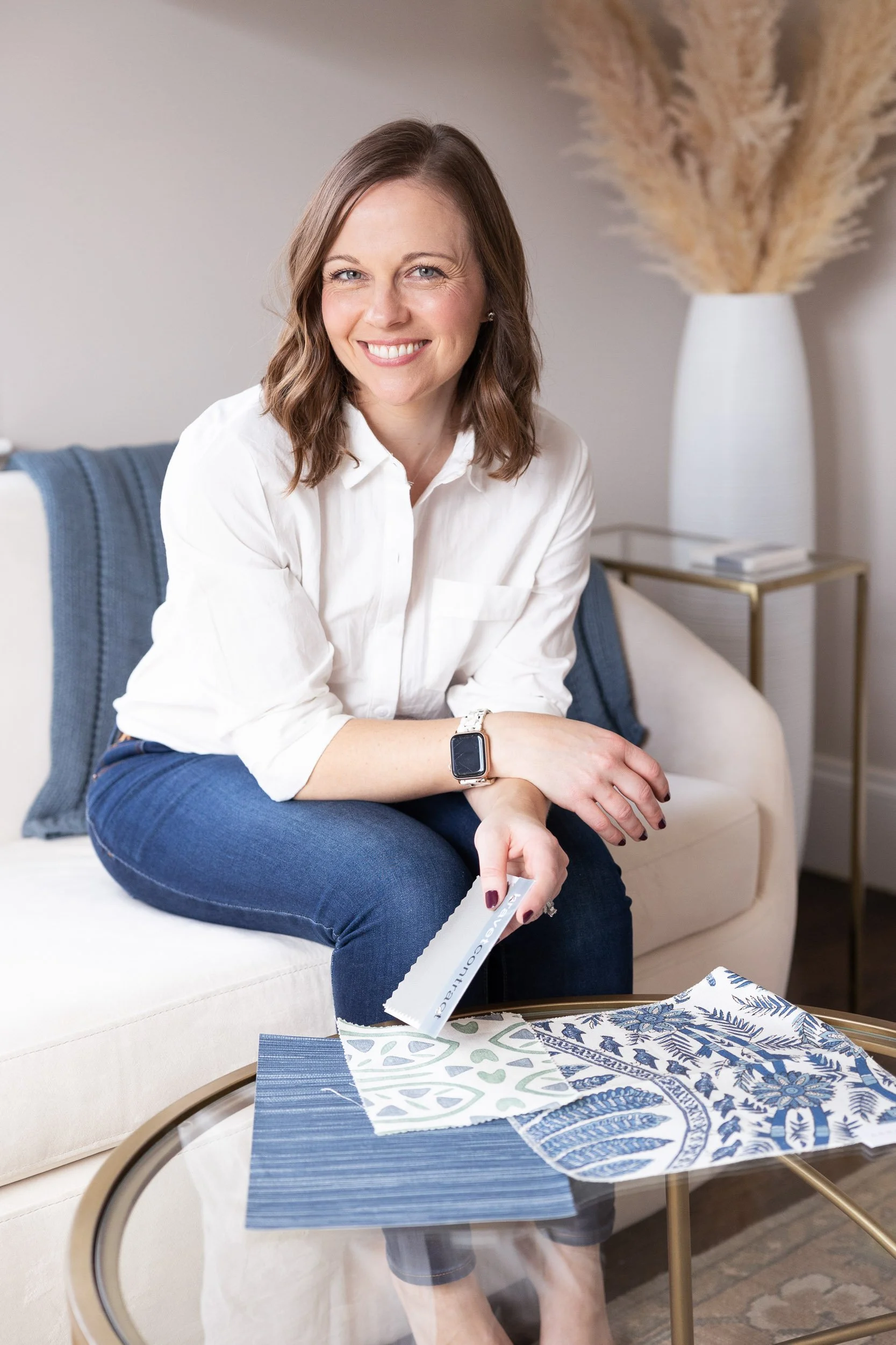 A Boston interior designer sitting on a beige sofa, smiling, wearing a white shirt, blue jeans, looking at fabric samples on a glass coffee table in a modern living room.