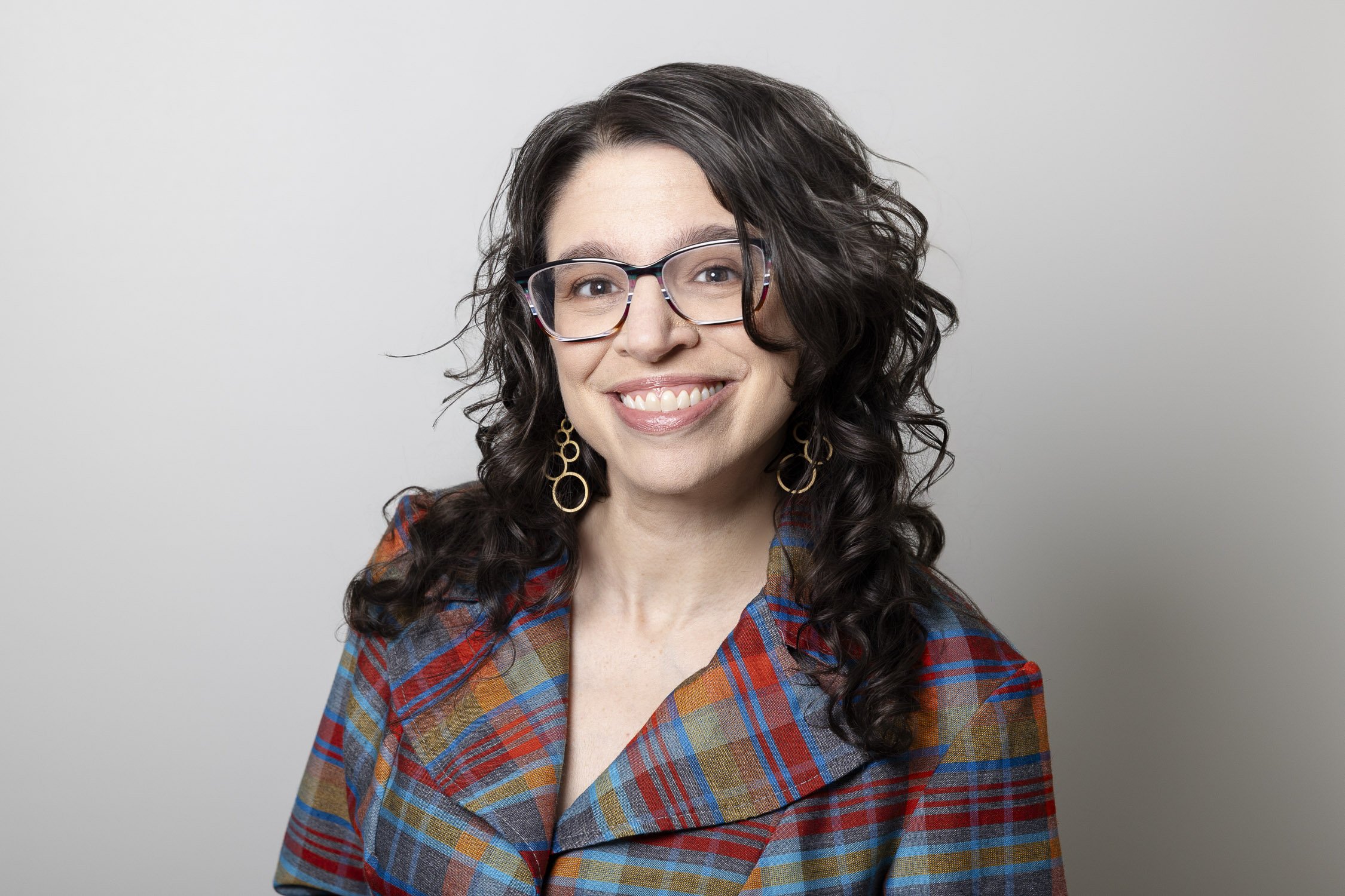 A woman with curly dark hair, wearing glasses, gold hoop earrings, and a plaid shirt, smiling at the camera against a plain background.