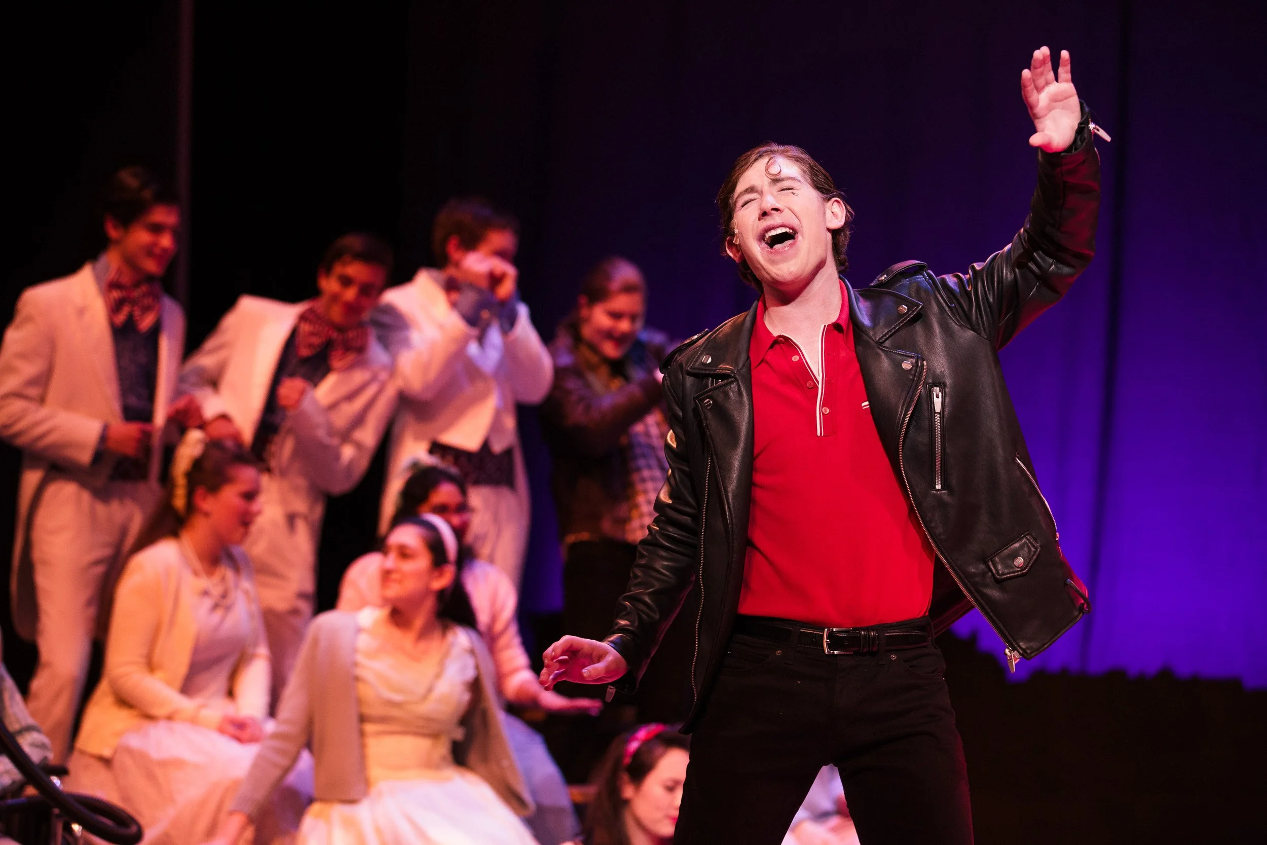 A male performer singing passionately on stage with a group of young people in the background, during a theatrical production at a private school in Boston, Massachusetts..