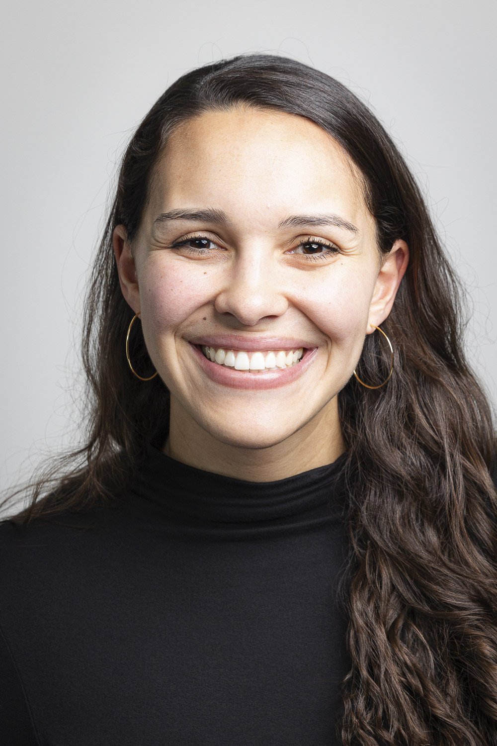 Close-up portrait of a woman with long curly brown hair, wearing black turtleneck, with a big smile against a gray background.