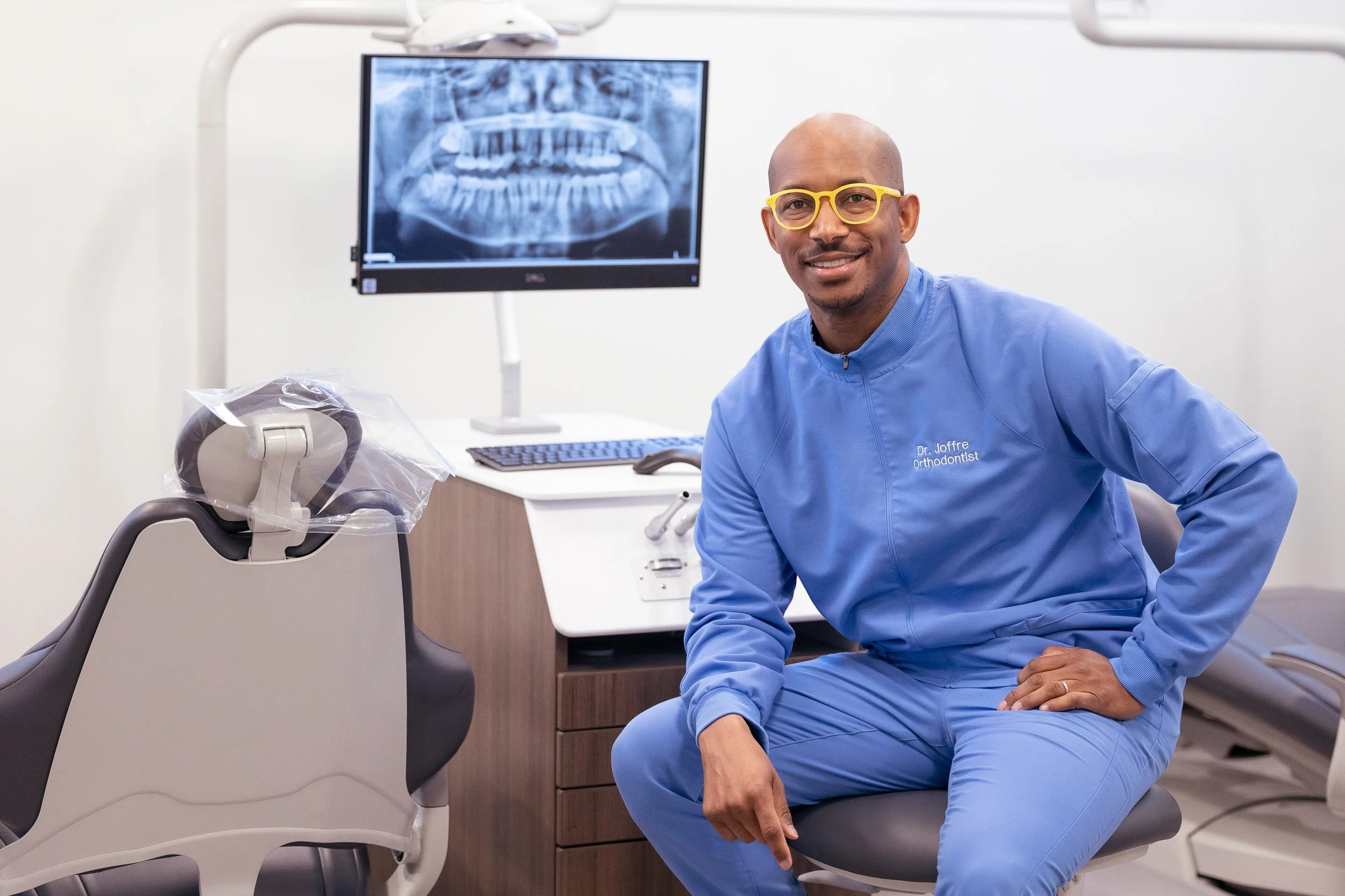 A male orthodontist in blue scrubs and yellow glasses sitting in a dental office with an X-ray monitor behind him. 